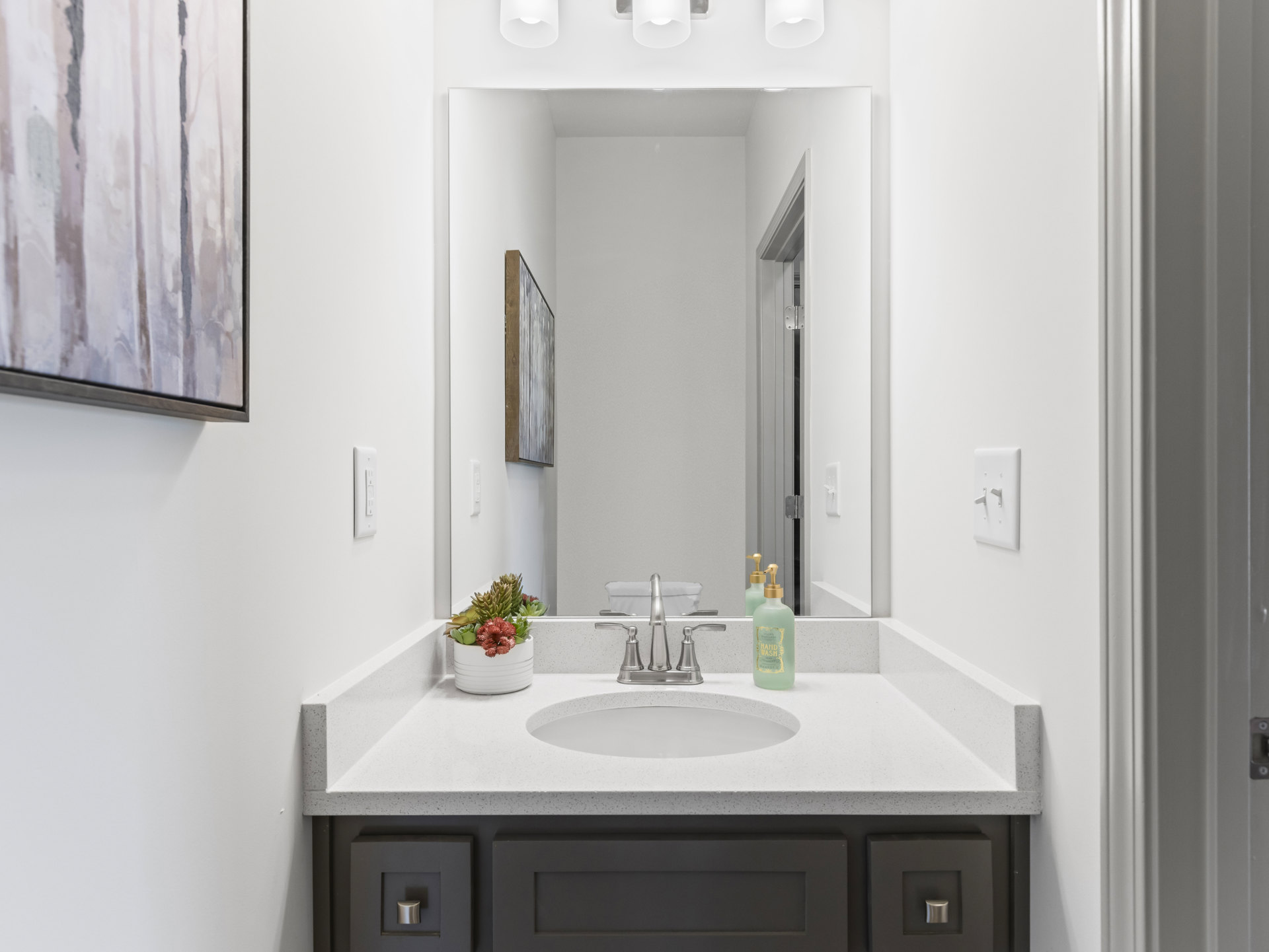 Modern bathroom with white walls, rectangular mirror above a sleek sink, chrome faucet, potted plant, bottle of hand wash on the countertop, and trees visible through a window in