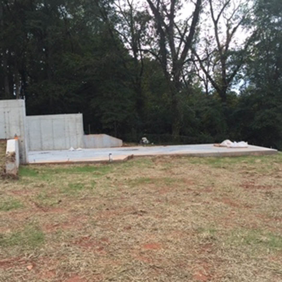 Modern concrete home set in a grassy field, surrounded by mature trees, with a paved road and white post visible in the foreground