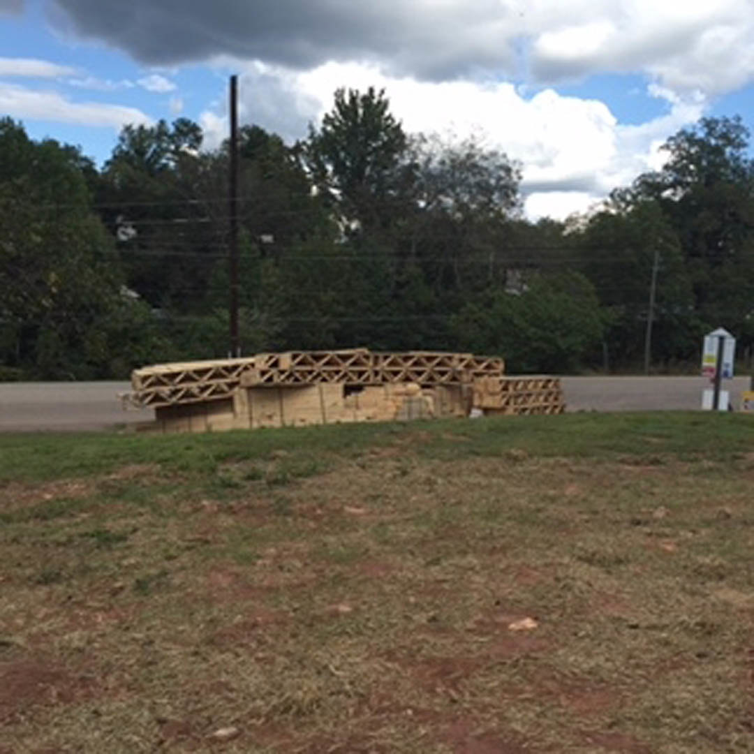 Stacked lumber beside a paved road, grassy field and partially constructed wooden-framed building in background, scattered trees under cloudy sky