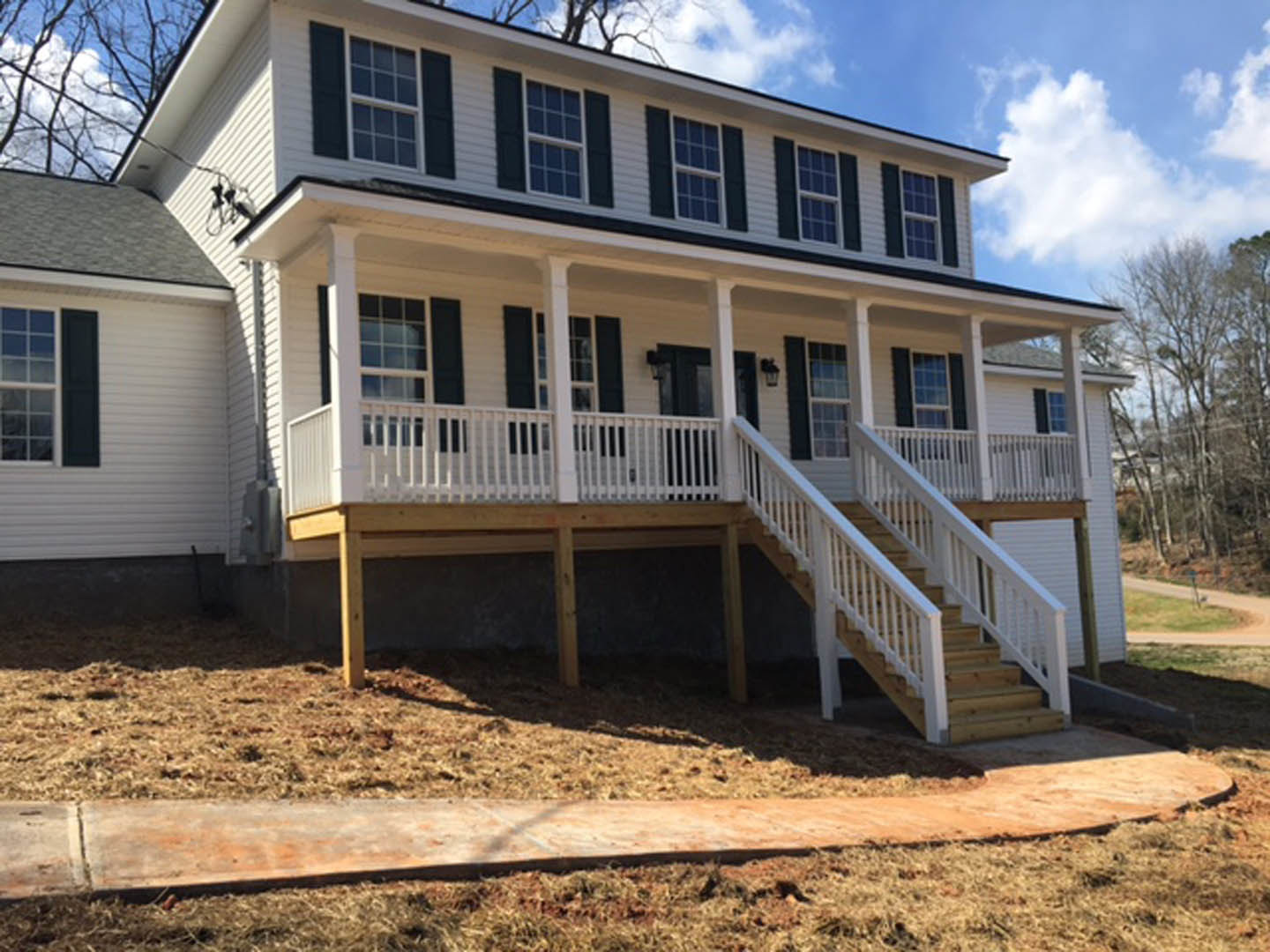 Front porch with white railings and stairs, concrete slab walkway, light-colored siding, windows, trees in background, blue sky with scattered clouds