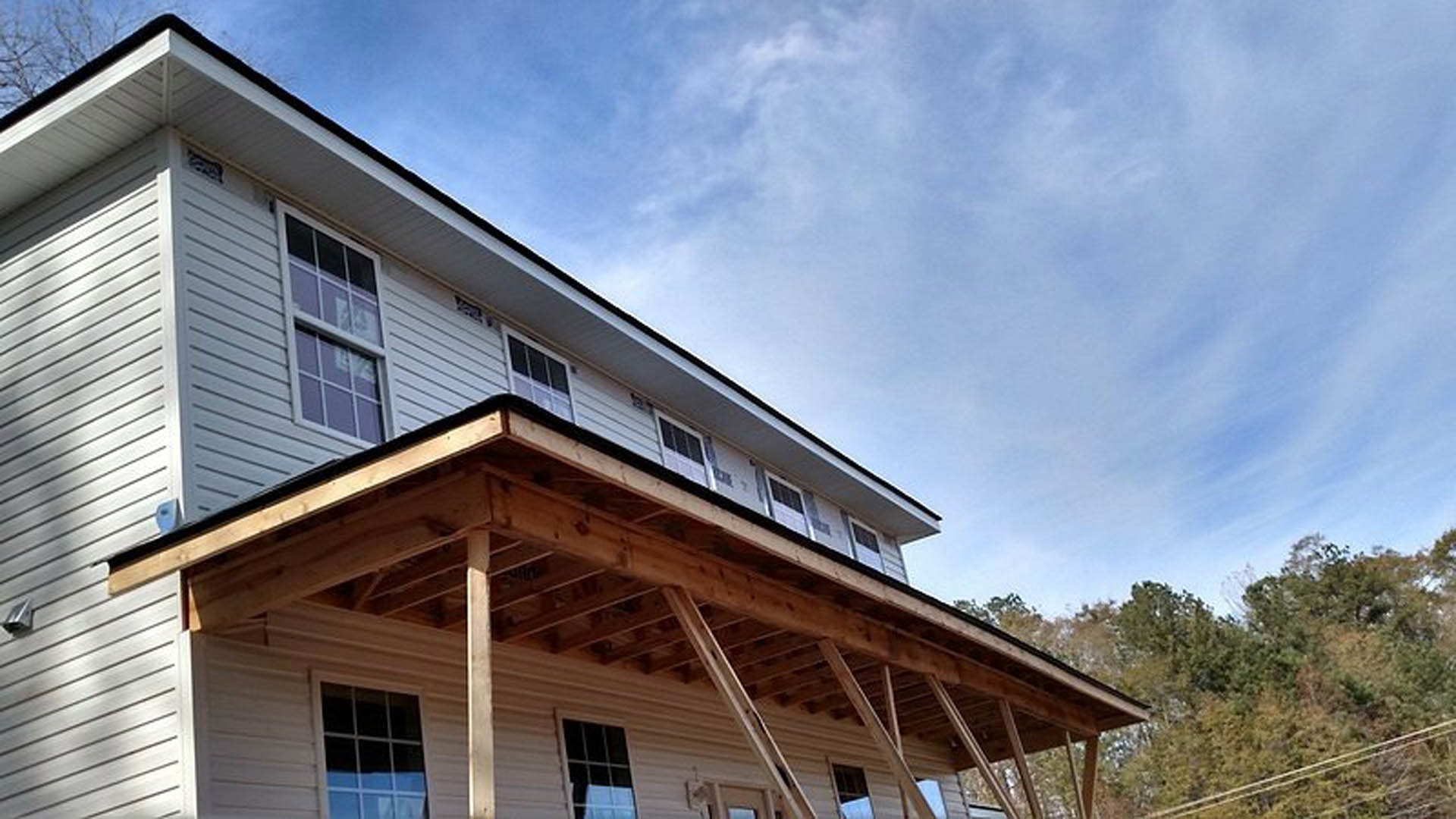 Wooden porch with white railings, gray siding, large windows with white curtains, gabled roof, and mature trees in the background
