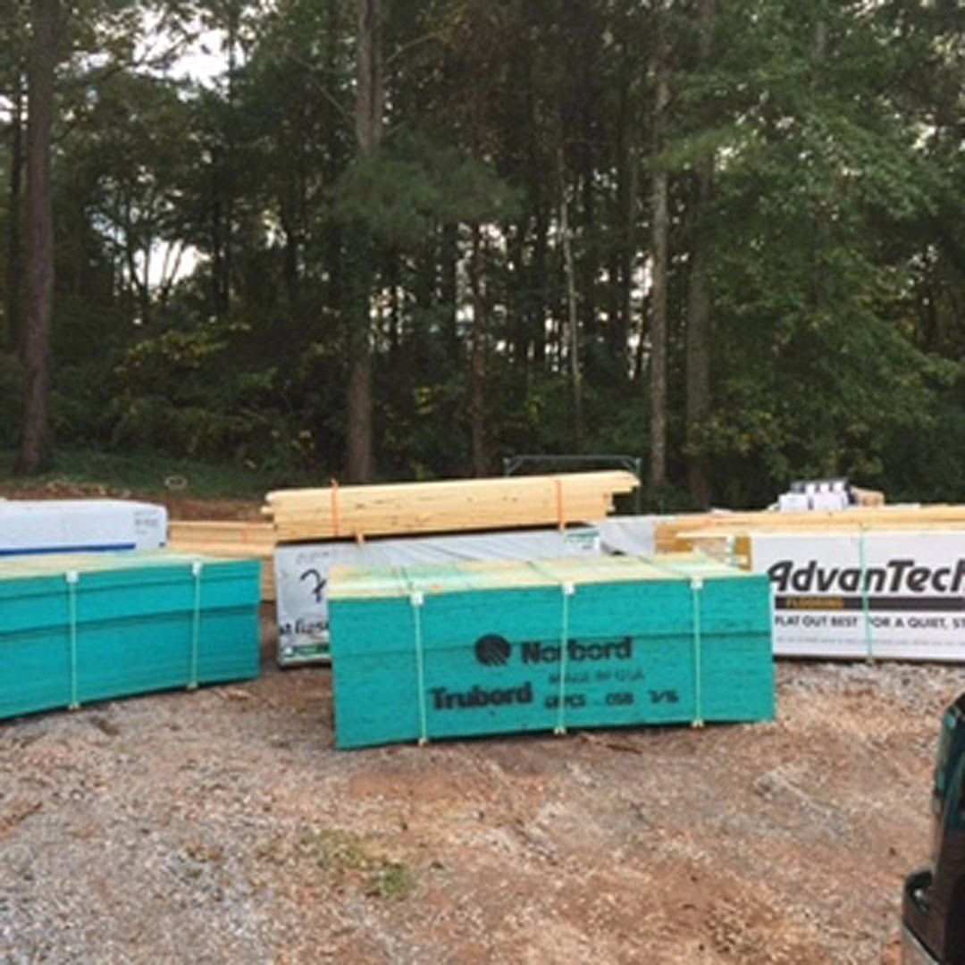 Stacked lumber and wood framing materials on bare ground outdoors, surrounded by trees, with a blue box labeled in black text nearby.
