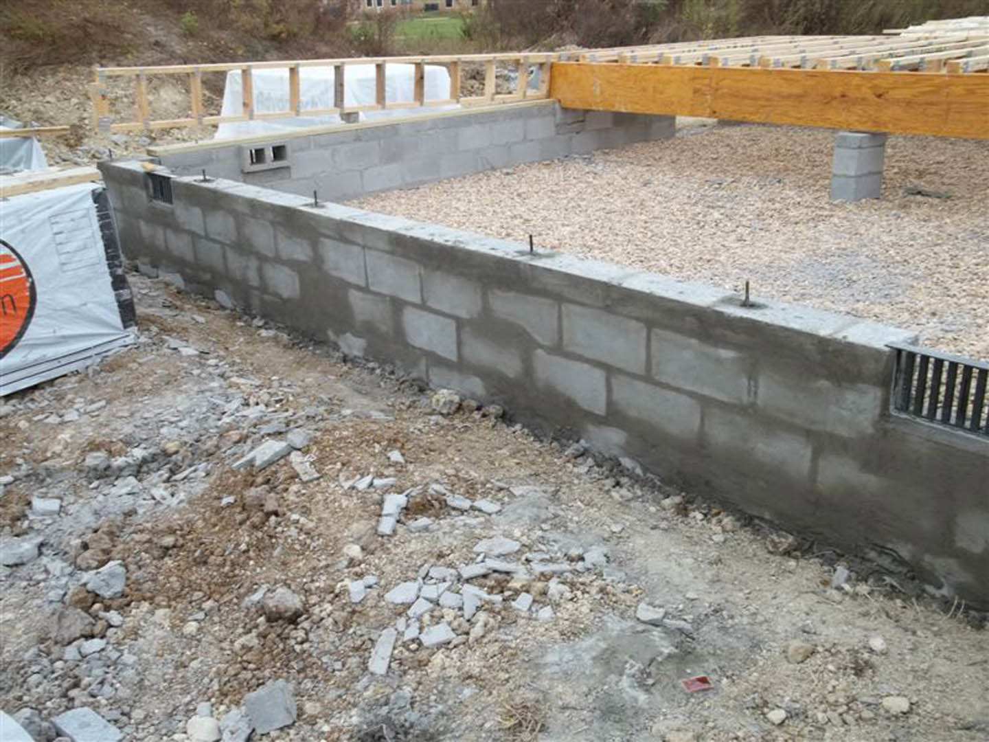 Concrete foundation under construction with exposed gravel, rocks, white tarp, orange construction equipment, wooden boards, and temporary fencing in an outdoor setting.