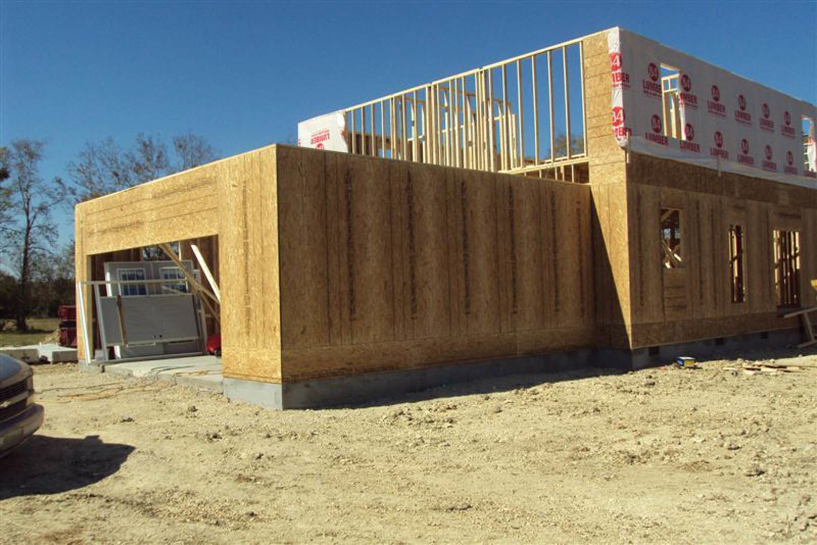 Wood-framed custom home under construction with exposed metal beams, dirt field in foreground, several workers on site, white wall featuring red logo, trees and blue sky in