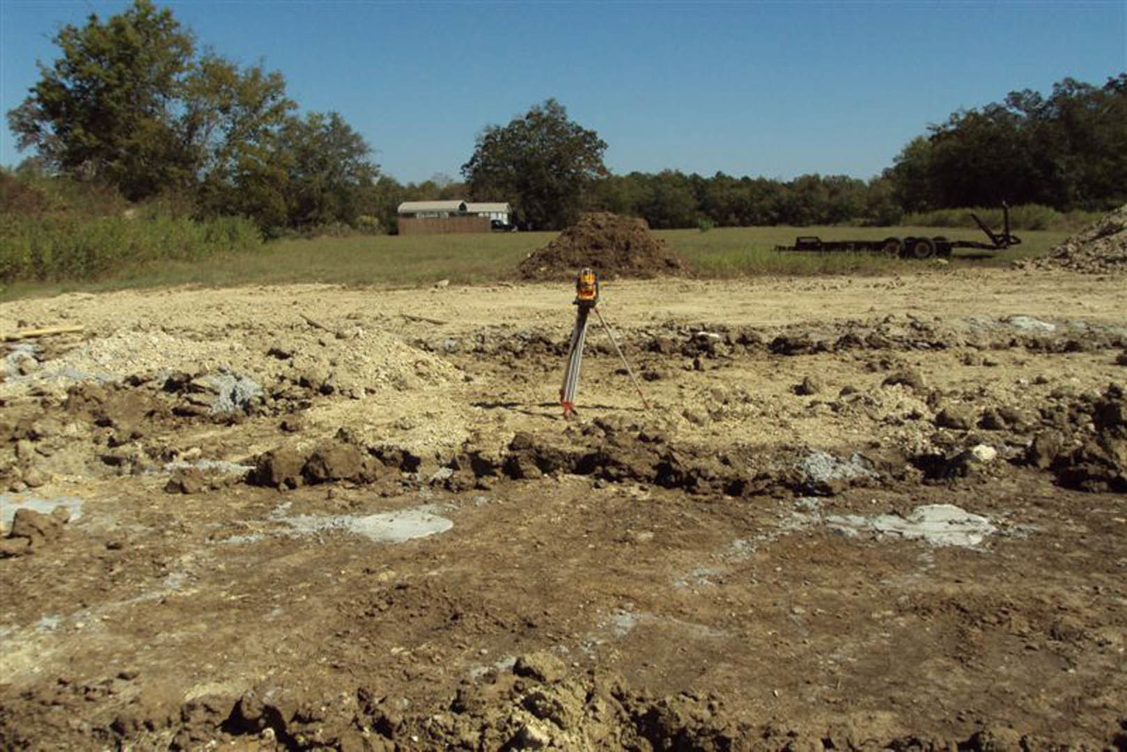 Land surveyor equipment set up in grassy field with muddy soil, distant building visible, tree and blue sky in background