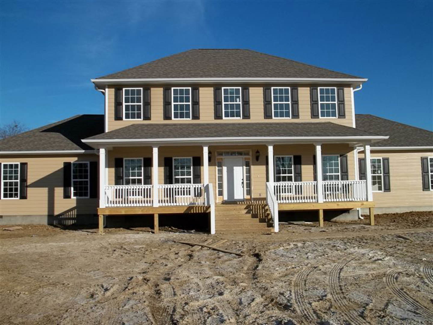 White house under construction featuring a front porch with white railings, glass-paneled door, black shuttered windows, and exposed dirt ground with wooden beam.