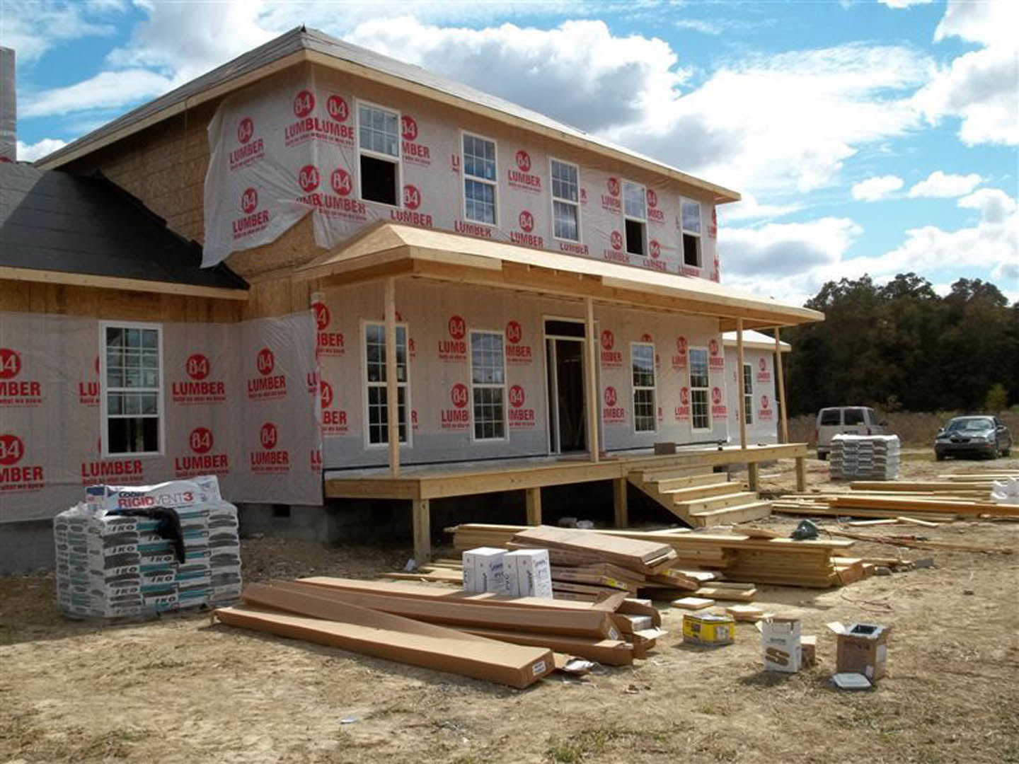 Wood-framed house under construction with stacked lumber, cement blocks, and boxes in foreground; tall tree and cloudy sky in background; partially installed window visible