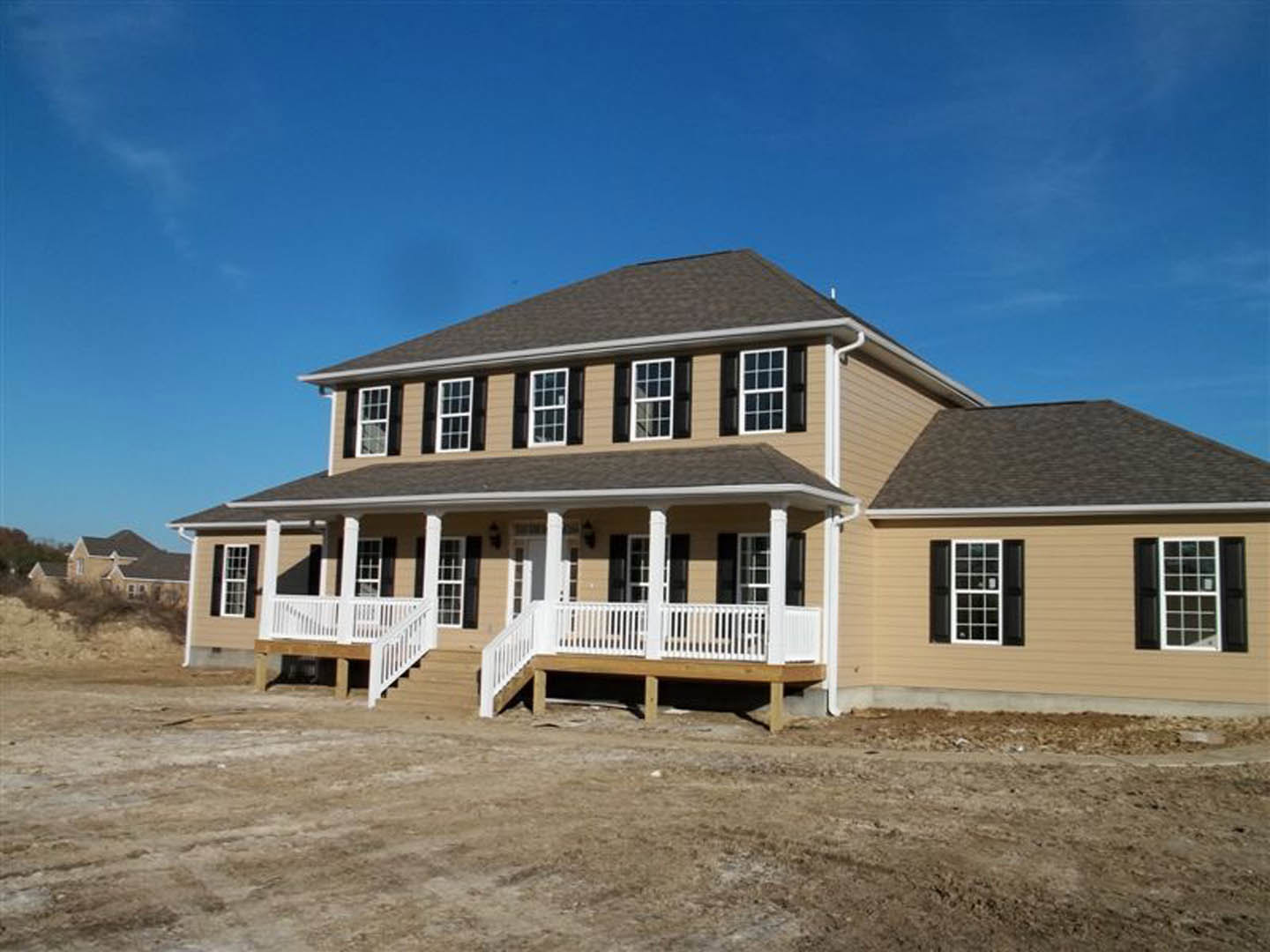 Partially built house with white deck railing, porch, white door featuring black square windows, brown dirt ground, and blue sky