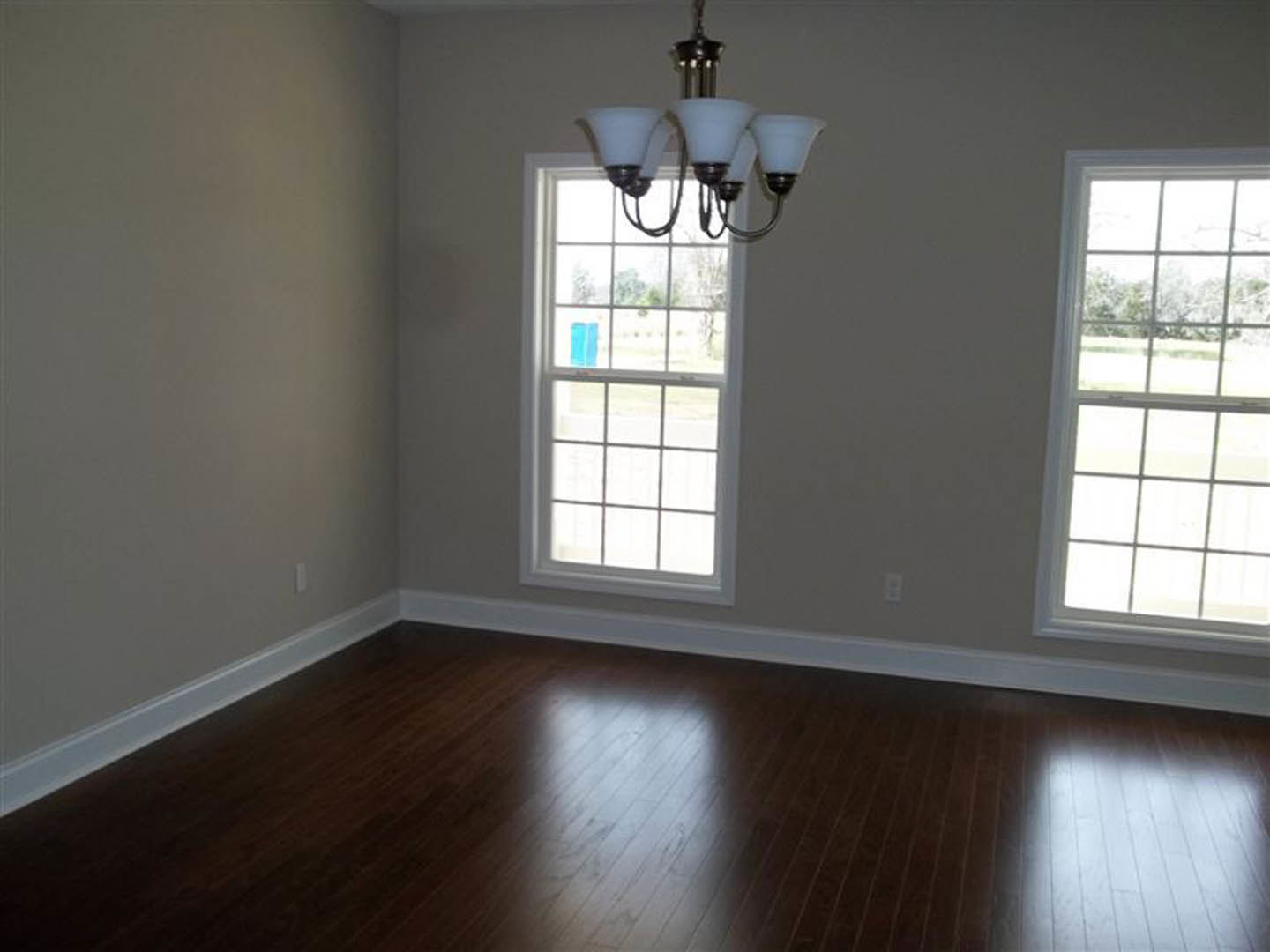 Chandelier hanging from a plaster ceiling above hardwood floors, multiple-paned windows with blinds framing a view of a grassy field, light reflecting across laminate flooring.