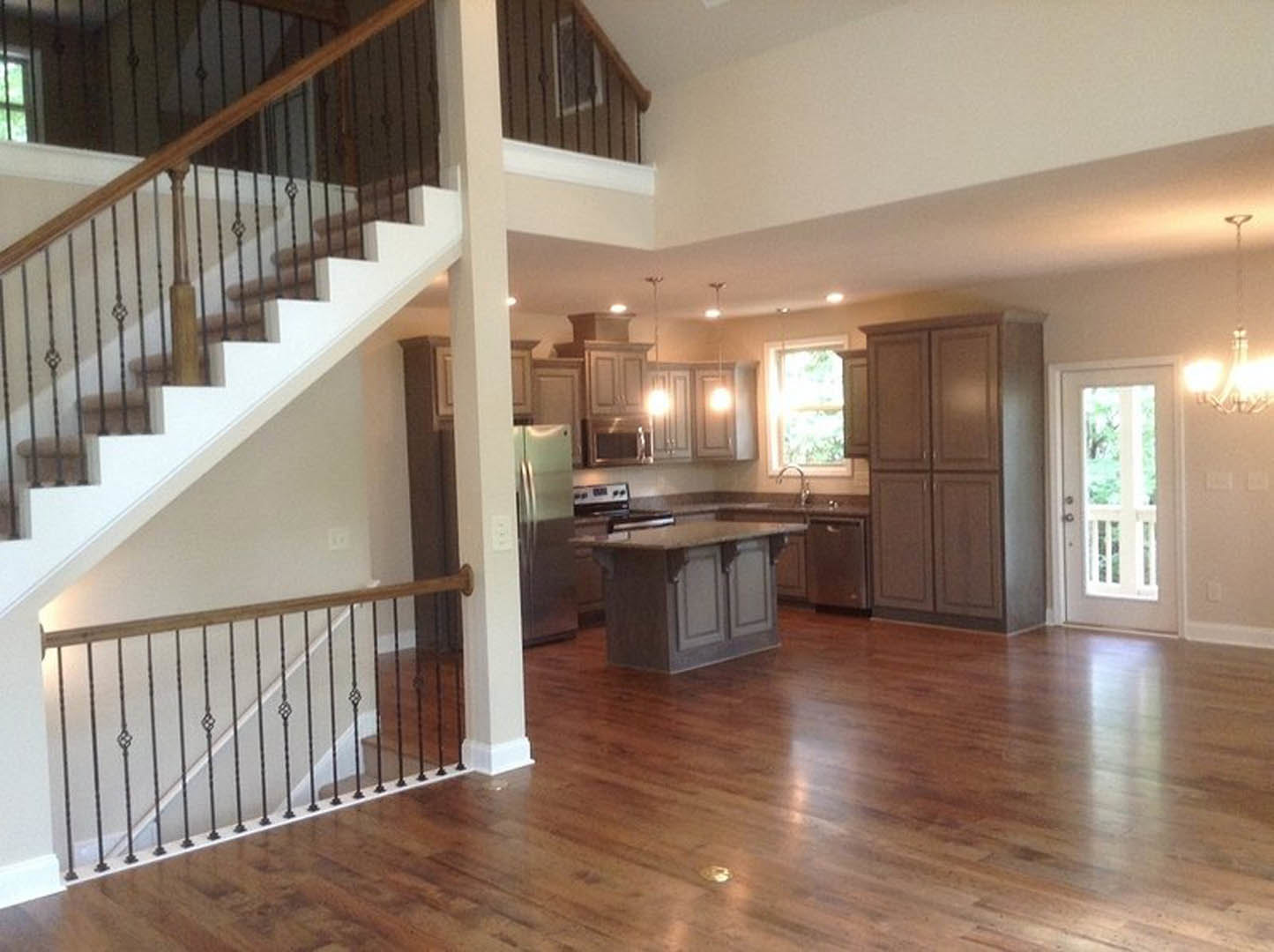Open kitchen with marble island, hardwood floors, white cabinetry, adjacent staircase featuring white railing and glass-paneled door, white column separating spaces.