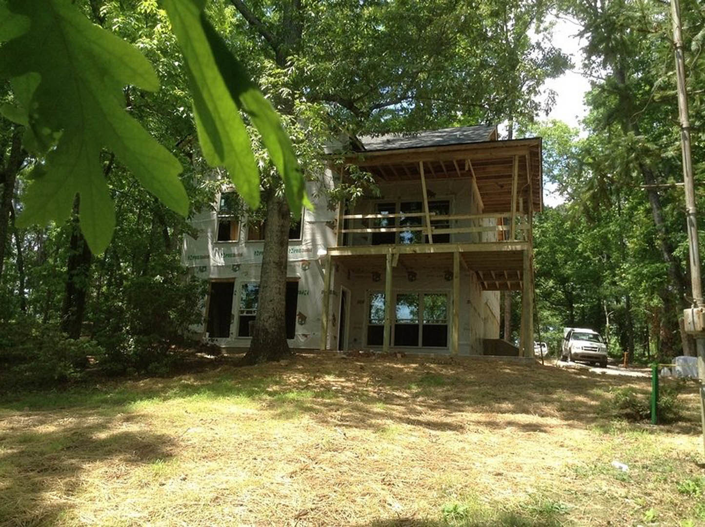 Wood-sided house with covered front porch, mature tree on grassy lawn, blue sky overhead, car parked on street, construction materials visible near entry.