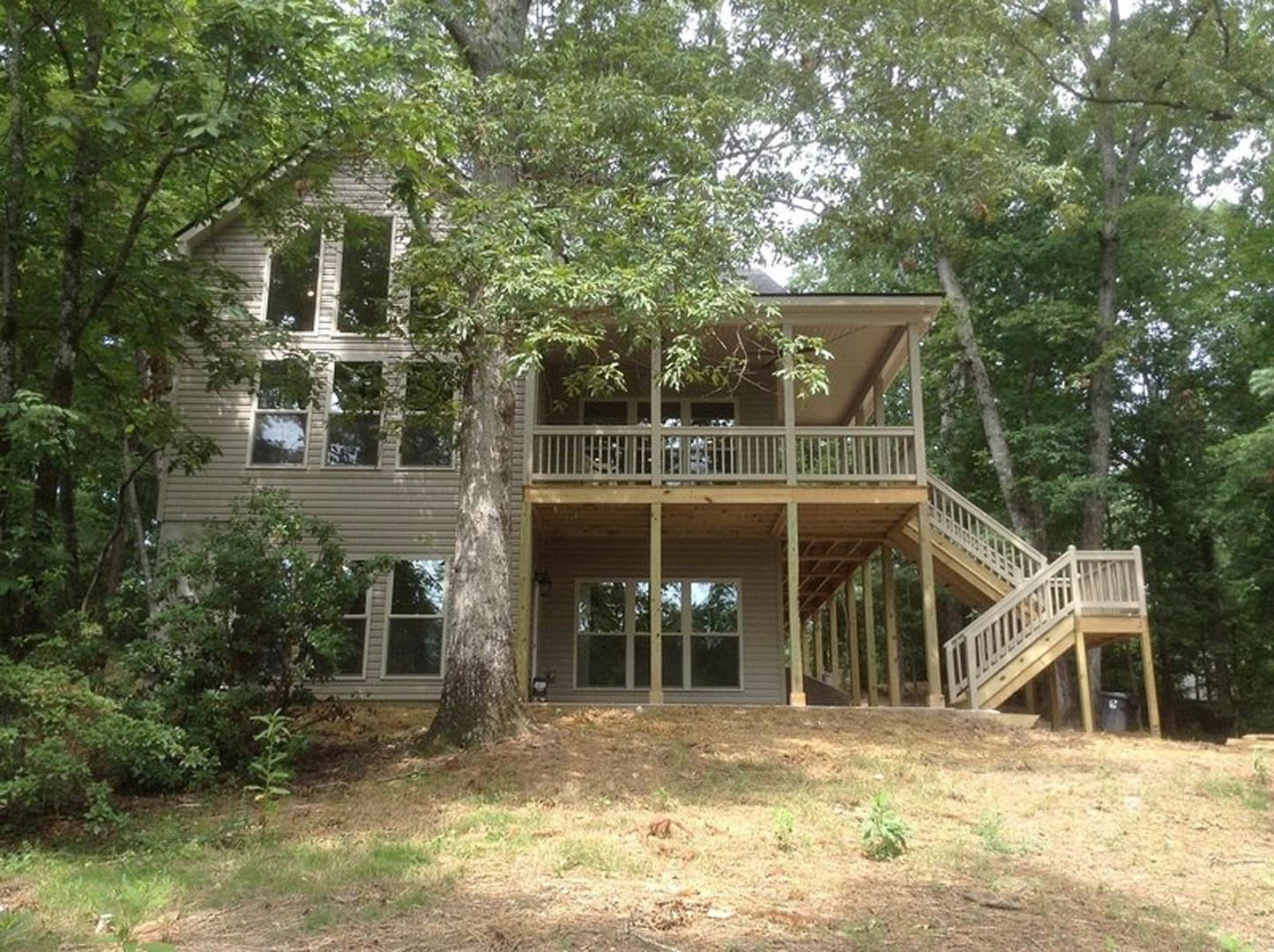Two-story wood-sided house with elevated deck and staircase, surrounded by tall trees and dense greenery