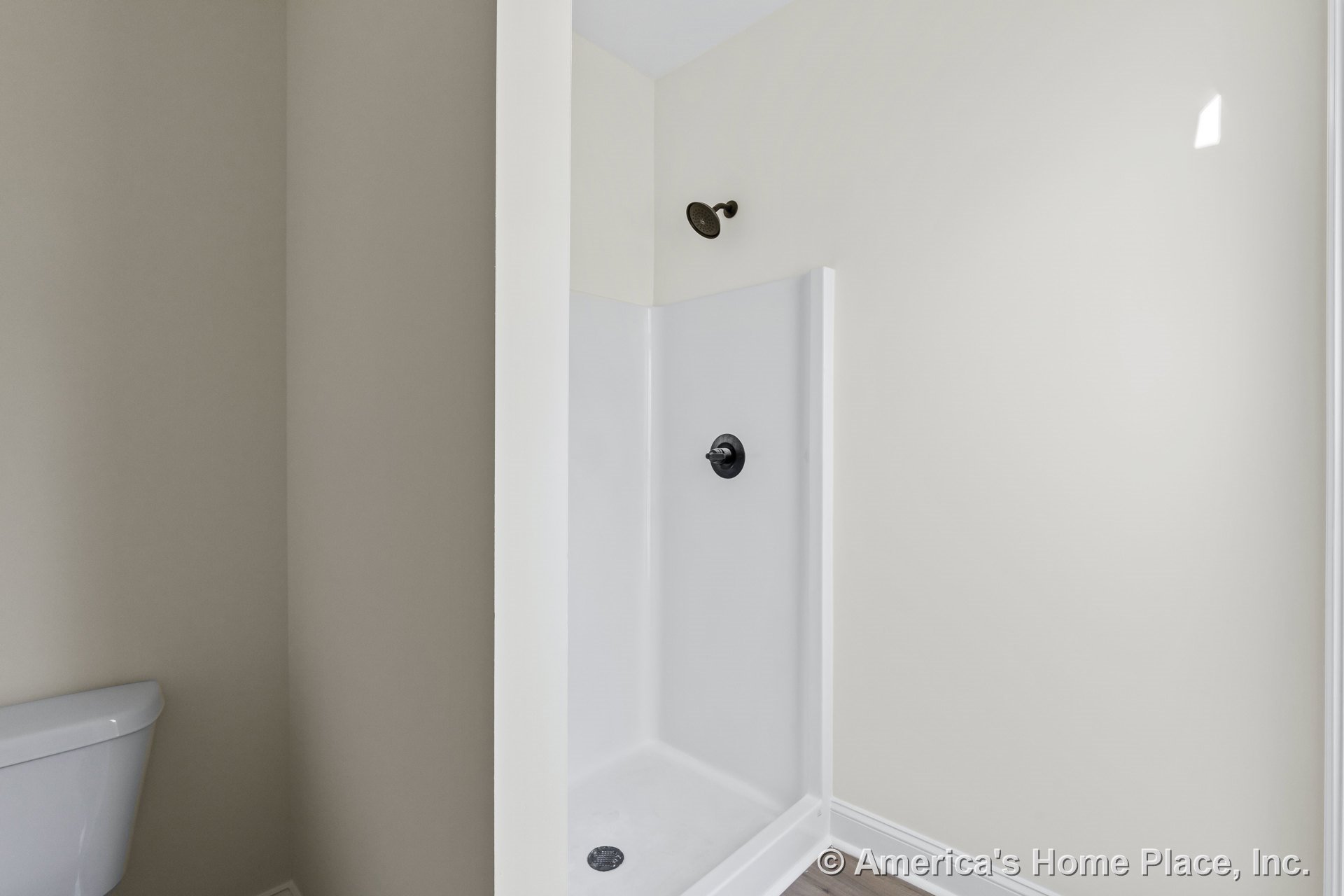 Walk-in shower enclosure with single showerhead, white surround, light-painted walls, partial toilet view, wood-look flooring, and minimal built-in trim.