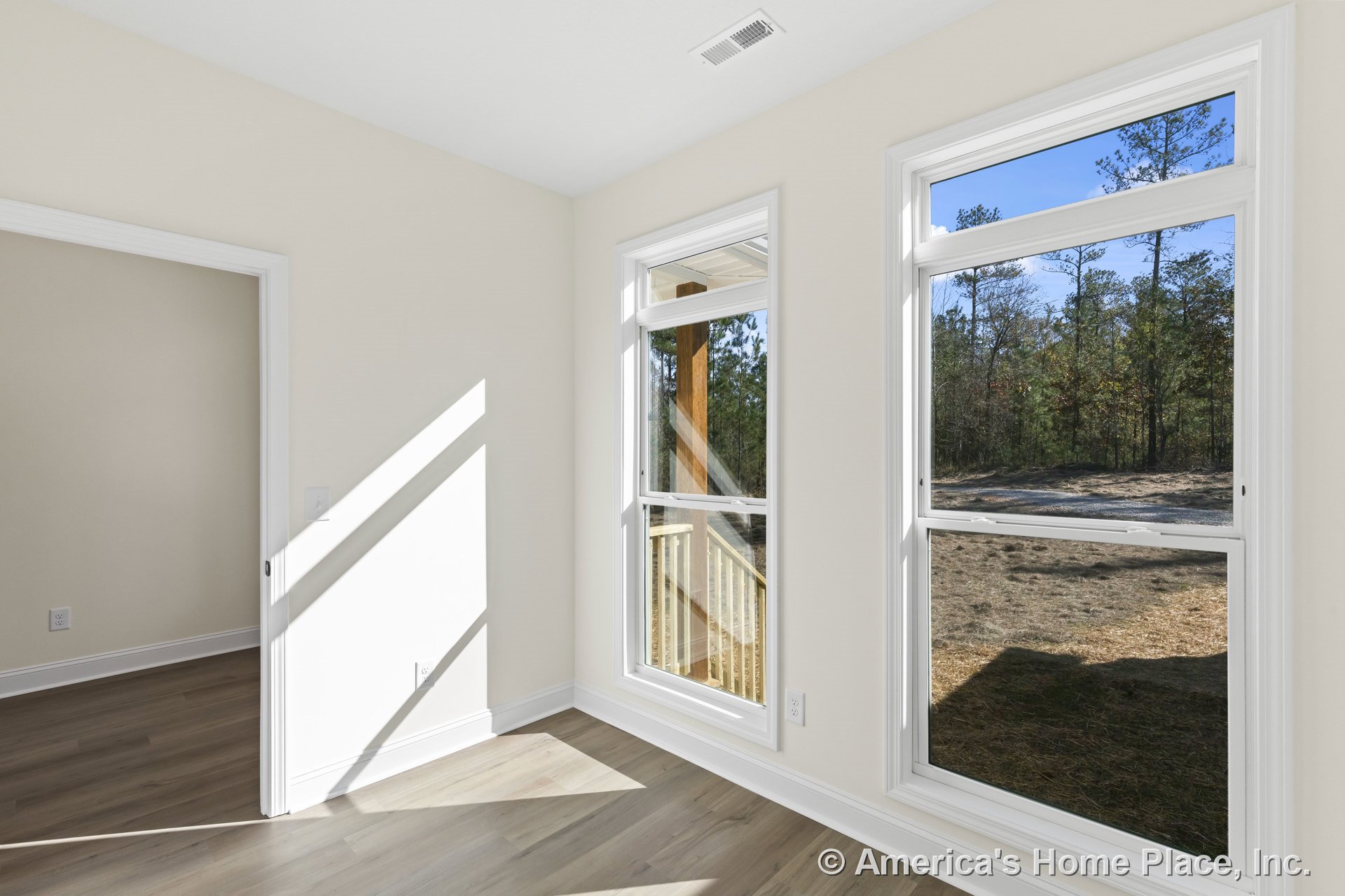 Corner of a bright interior room with large double-hung windows framed in white trim, light wood flooring, neutral wall color, baseboard molding, ceiling vent, and doorway leading