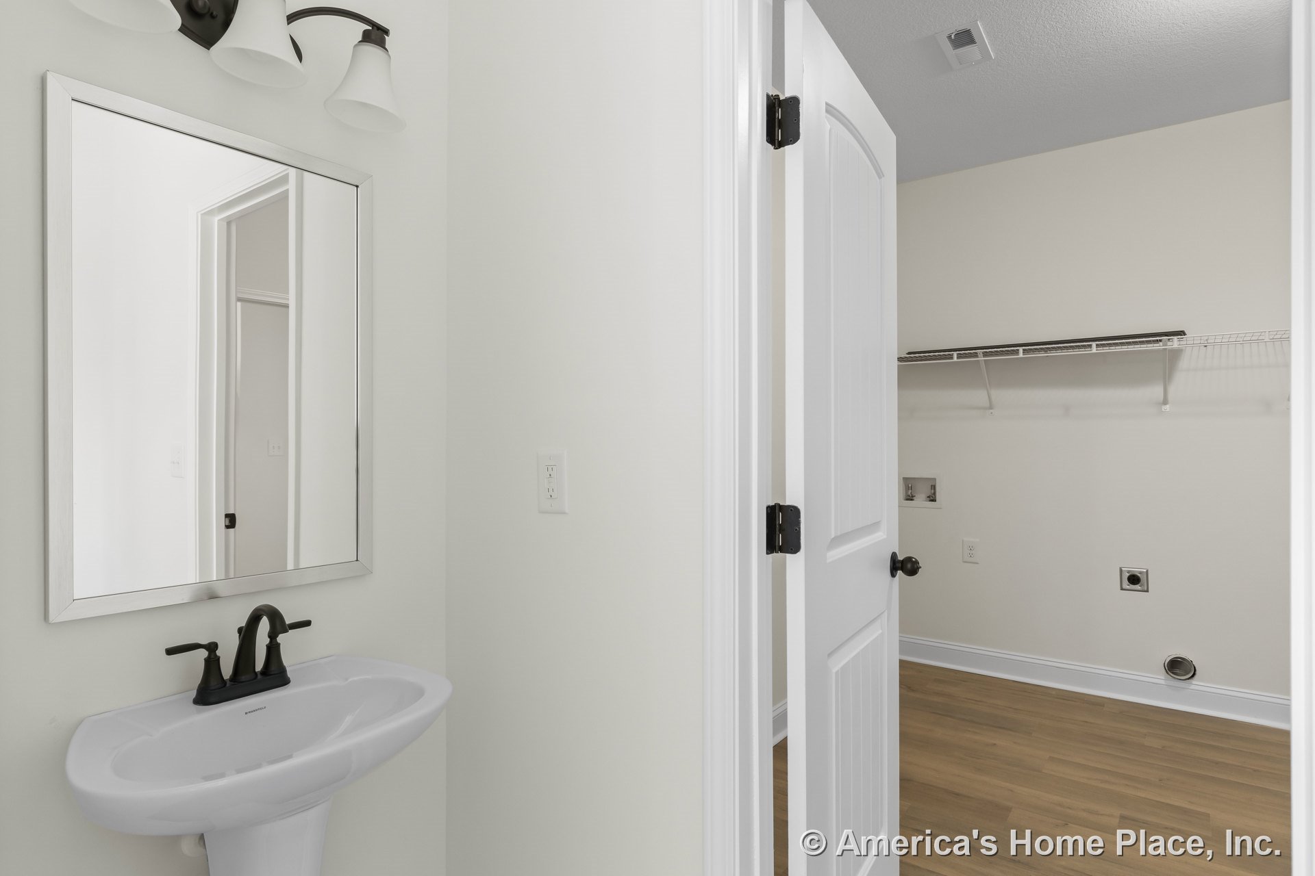 Laundry room with wire shelving and utility hookups next to bathroom featuring pedestal sink, wall-mounted mirror, three-light fixture, wood-look flooring, white trim, and neutral