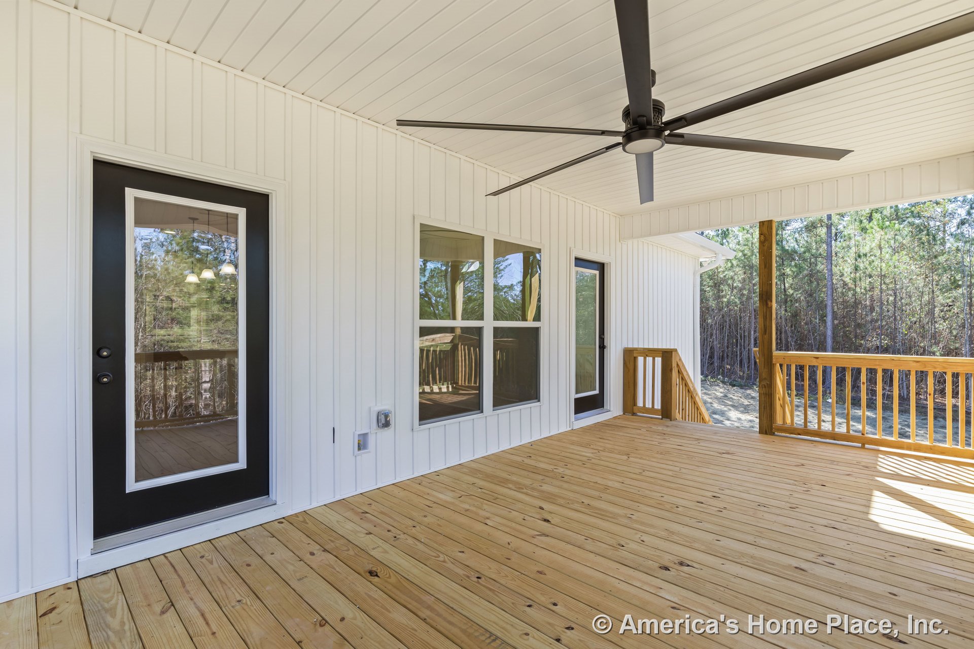 Covered porch with wood plank flooring, white vertical siding, paneled ceiling, large ceiling fan, black framed glass entry door, double window unit, wood railing, and white trim.