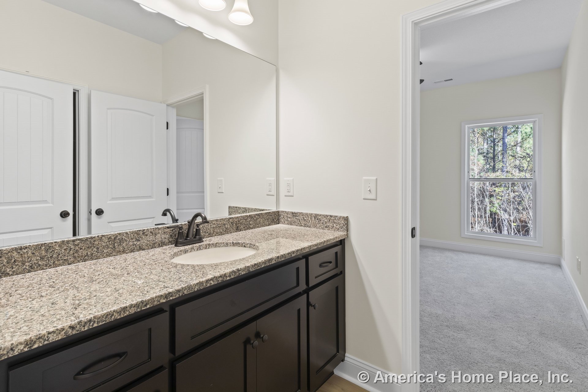 Granite countertop vanity with dark wood cabinets, large wall mirror, recessed lighting, white paneled doors, neutral wall paint, and view into carpeted adjacent room.