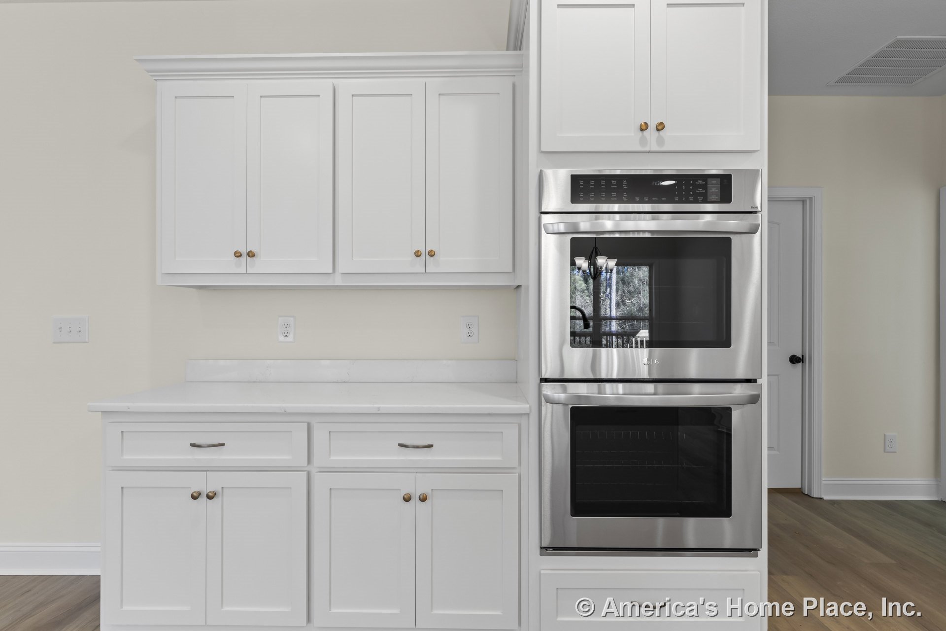 White shaker upper and lower cabinets with brushed nickel hardware, quartz countertops, built-in stainless steel double wall ovens, light wood flooring, neutral painted walls, and