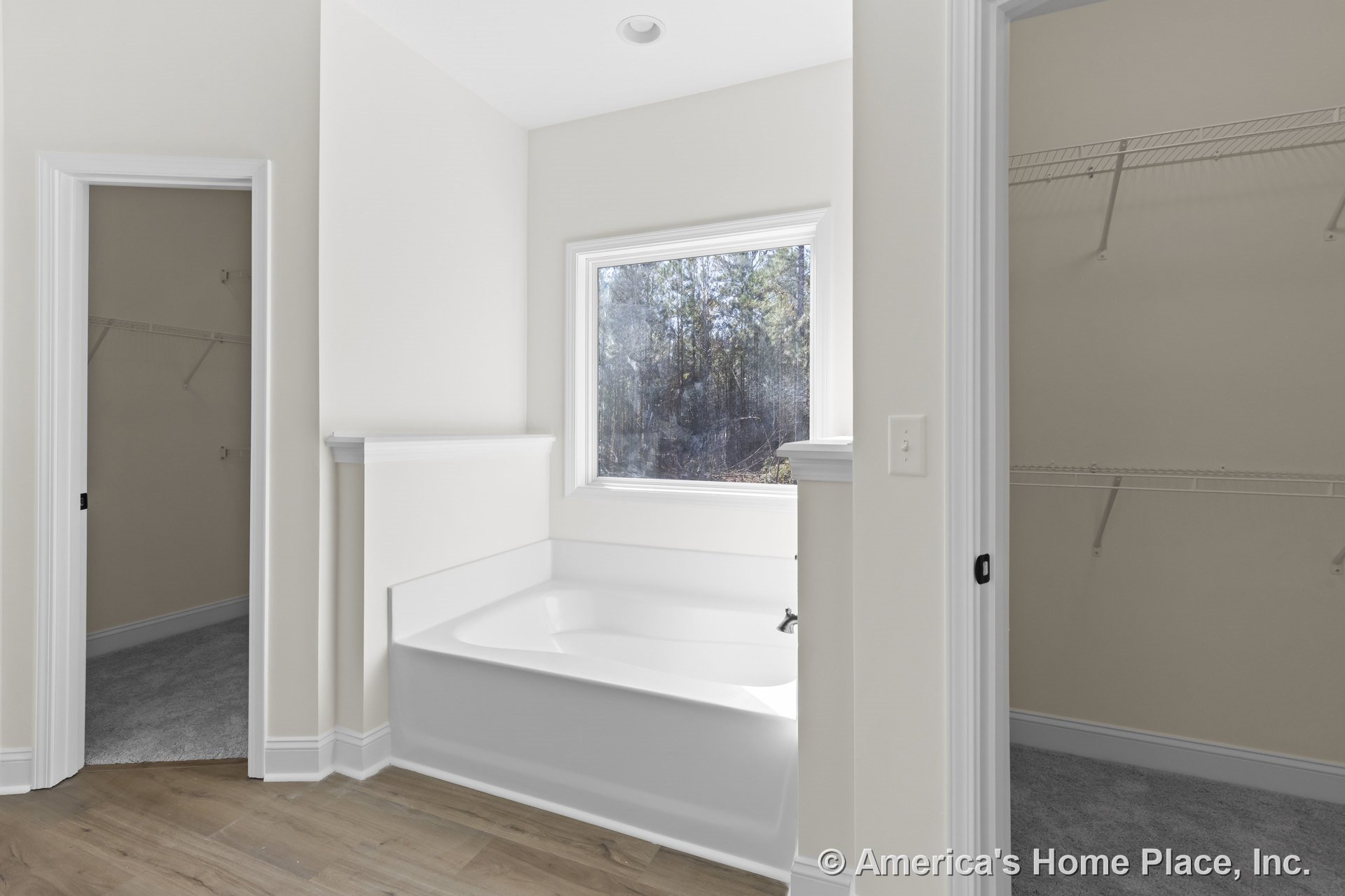 Built-in white soaking tub positioned beneath a large window with decorative trim, flanked by two walk-in closets featuring wire shelving; wood-look flooring, neutral wall color