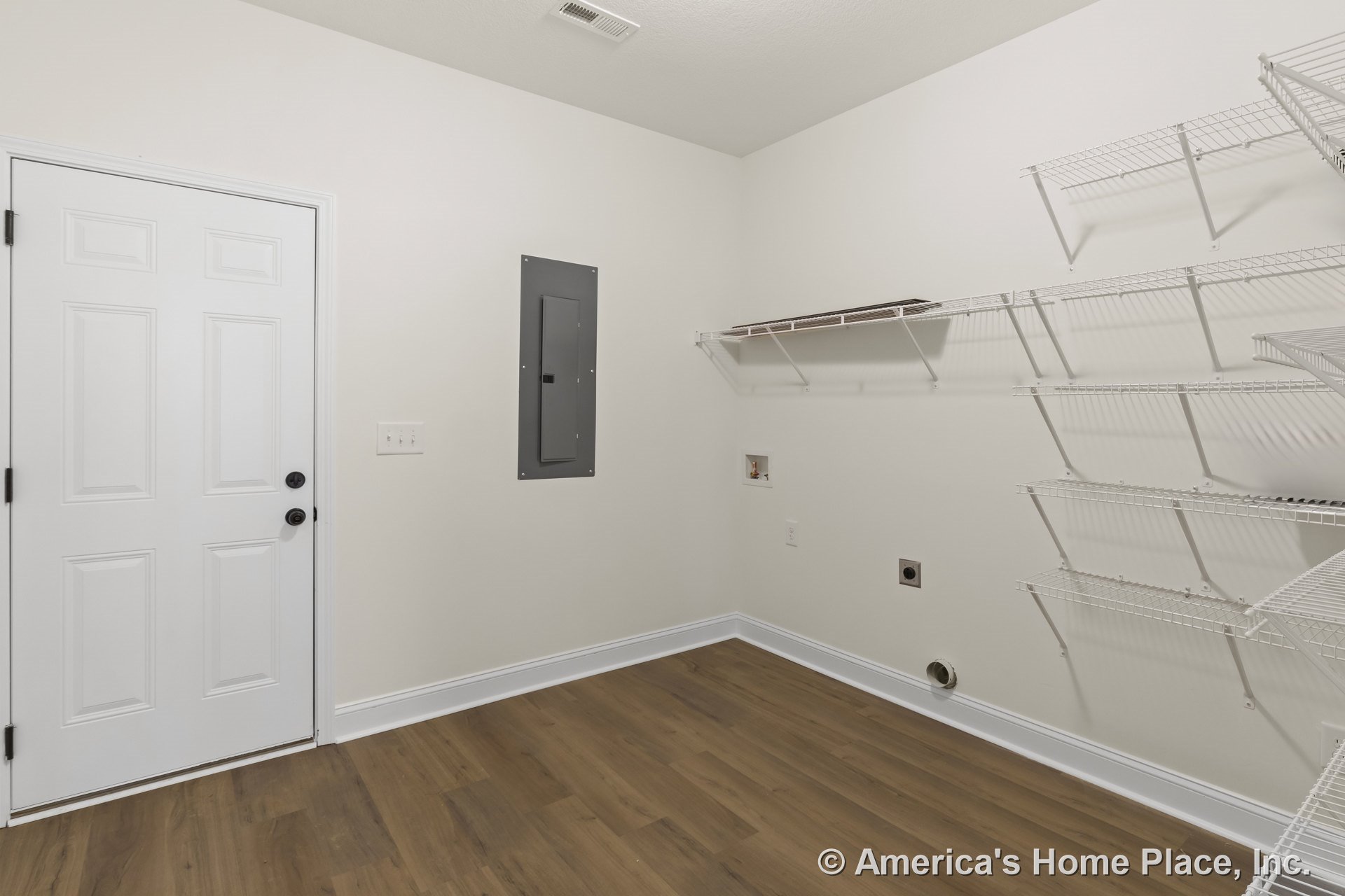 Laundry room with white wire shelving, utility hookups on wall, wood-look vinyl flooring, white paneled door, electrical panel, and white baseboards against light-colored walls.