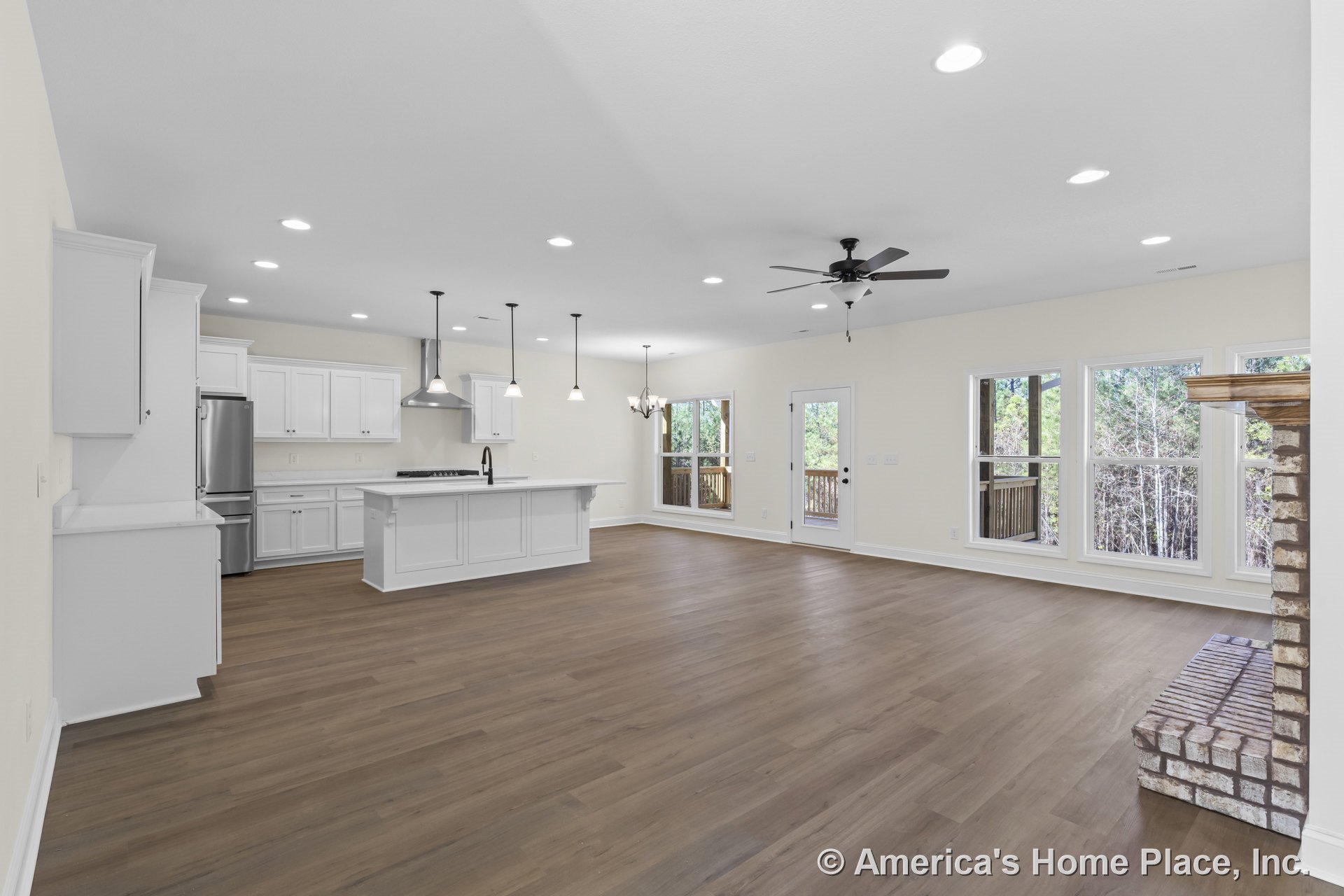 Open-concept living area with white kitchen cabinetry, central island with pendant lighting, stainless steel appliances, brick fireplace hearth, expansive window wall with trim