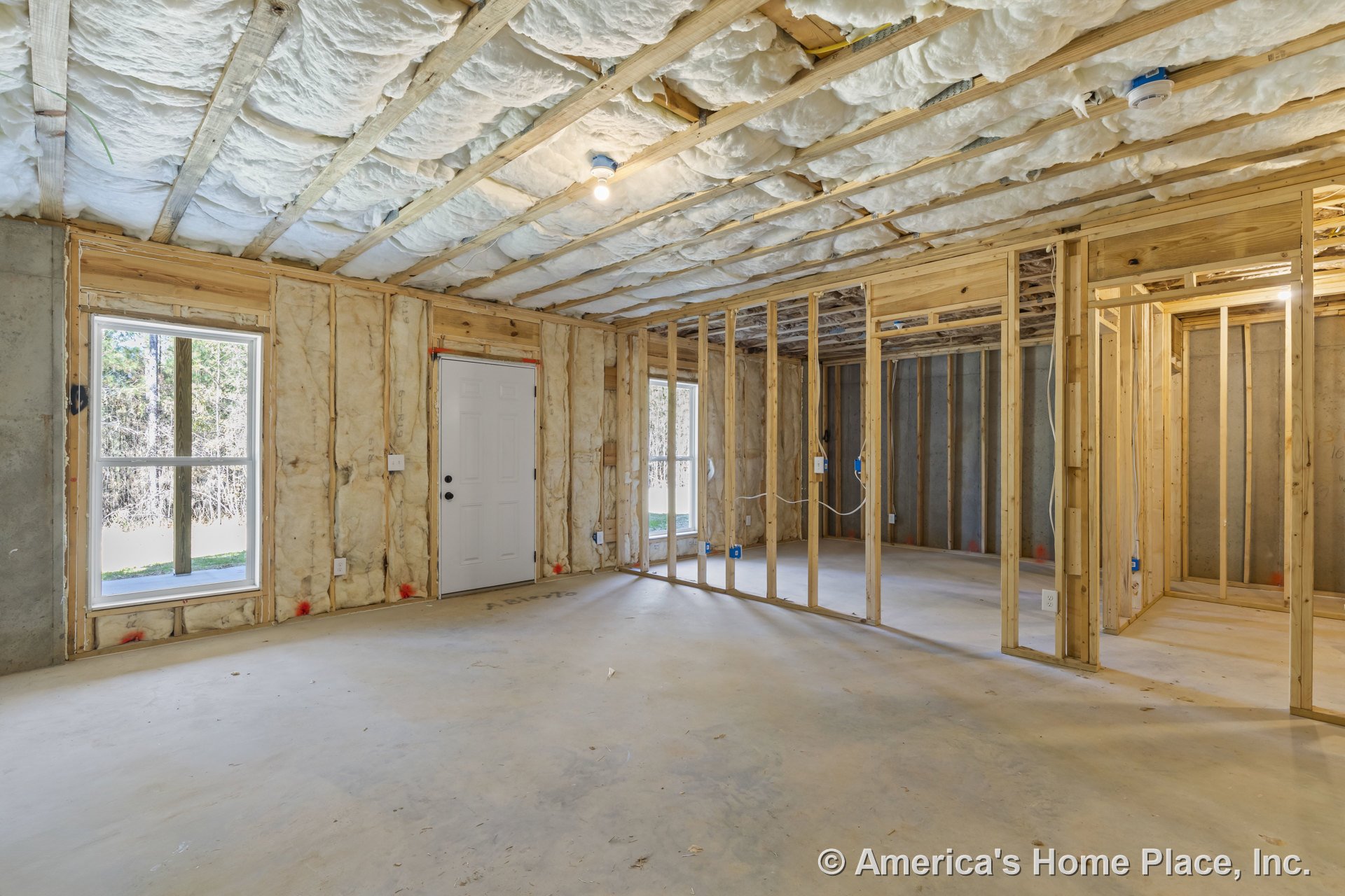 Unfinished basement with exposed wood framing, fiberglass insulation in ceiling, concrete slab floor, unfinished walls, white exterior door, double-pane windows, visible electrical