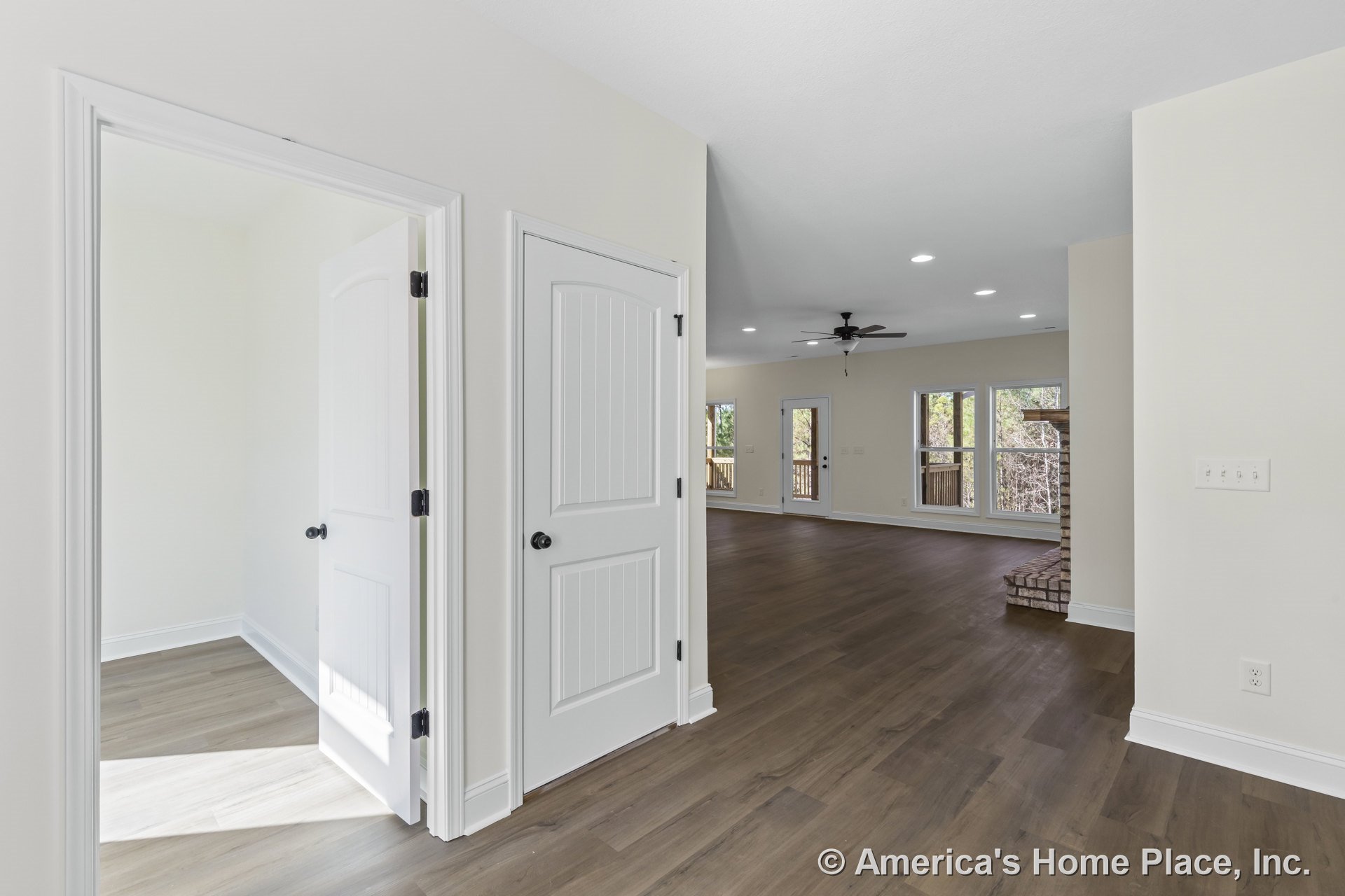 Open living area with wood-look plank flooring, neutral walls, white paneled interior doors, recessed ceiling lights, ceiling fan, large windows framed in white trim, and a brick