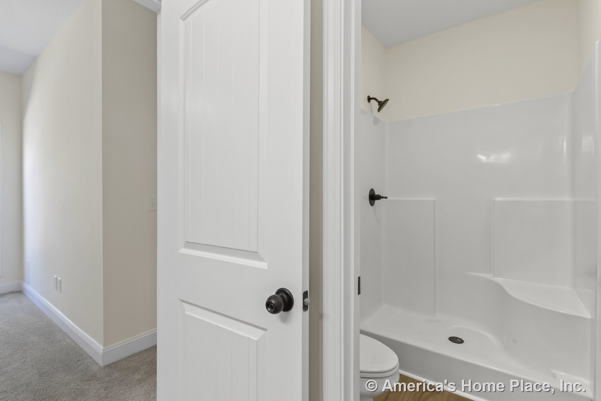 White fiberglass shower enclosure with built-in seat and bronze showerhead, light beige walls, wood-look flooring, and white paneled door.