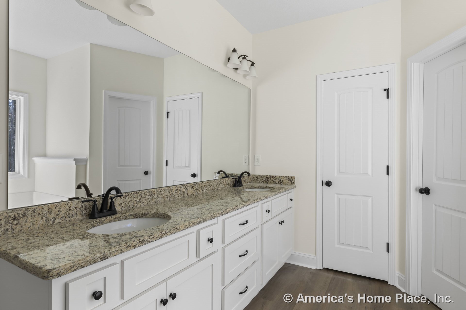 Long granite vanity with dual undermount sinks, white shaker cabinets, large wall mirror, matte black faucets, three-light vanity fixture, wood flooring, panel doors, and neutral