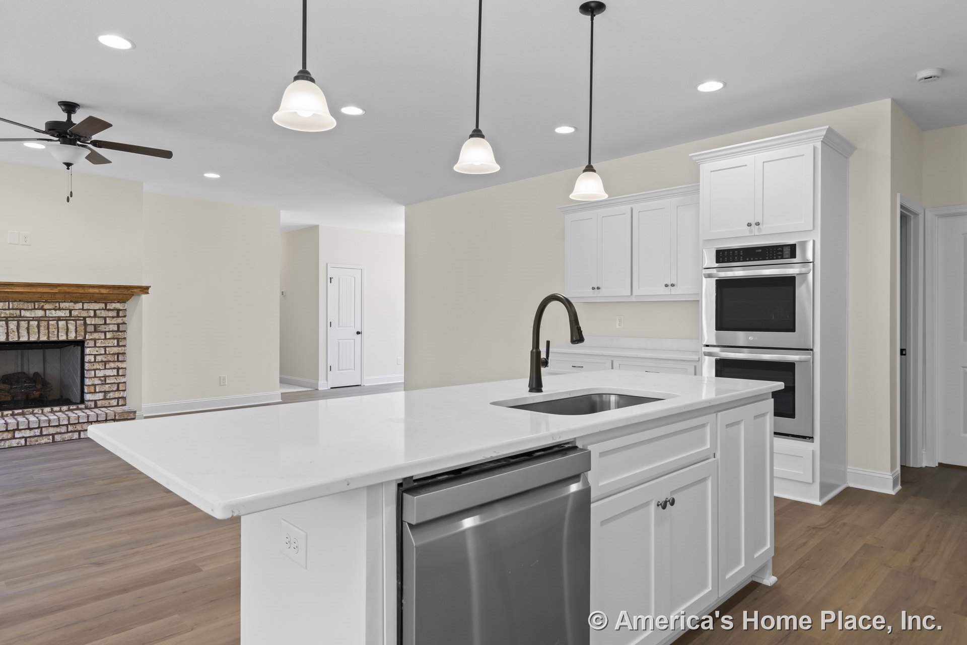 White shaker kitchen cabinets, quartz island with sink, stainless steel double oven, pendant lights above island, open floor plan leading to living area with brick fireplace and
