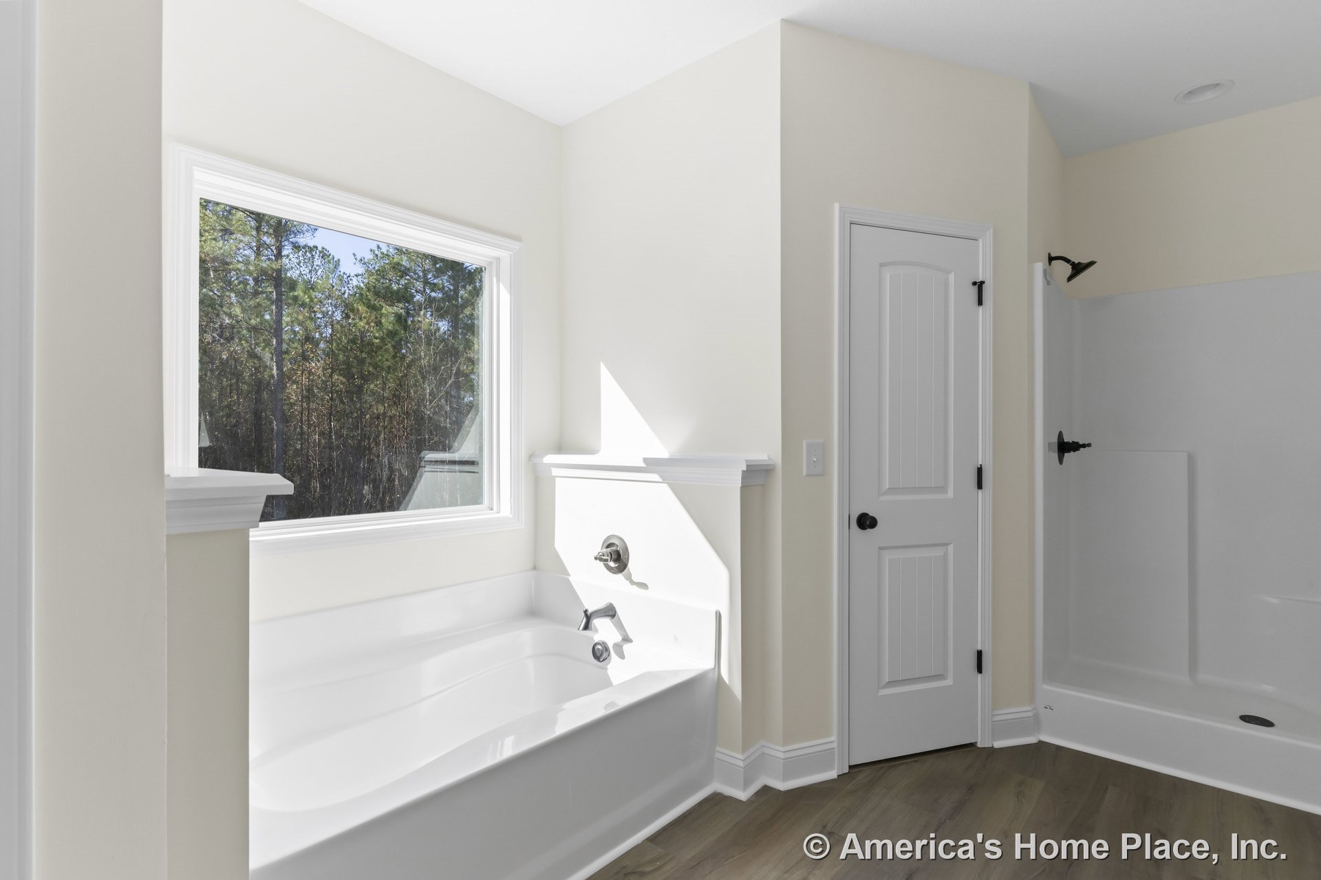 Built-in soaking tub with chrome faucet positioned under a large window with white trim, separate glass shower enclosure, wood-look flooring, paneled interior door, and neutral
