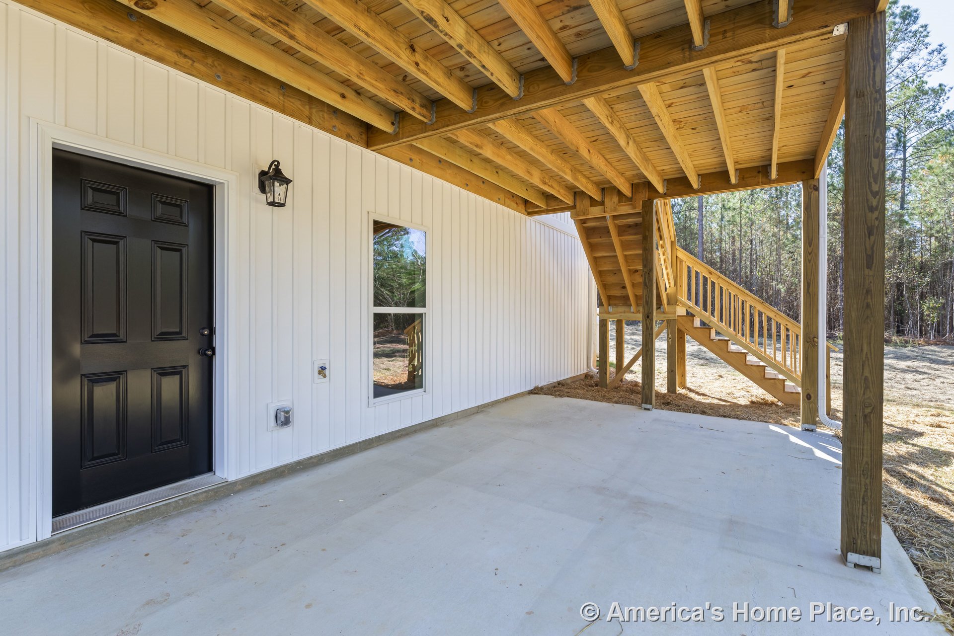Covered patio with concrete flooring, exposed wood beam ceiling, vertical white siding, black exterior door, single window, wooden staircase, wall-mounted light fixture, structural