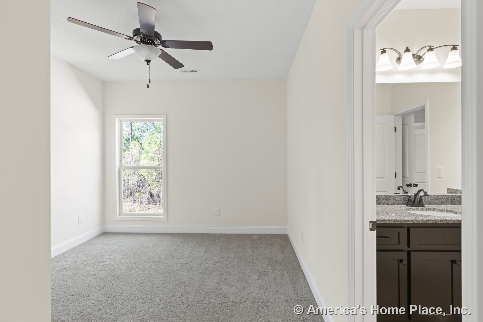 Carpeted bedroom with ceiling fan and large window with white trim, open doorway leading to bathroom featuring granite countertop, dark wood vanity cabinets, and modern light