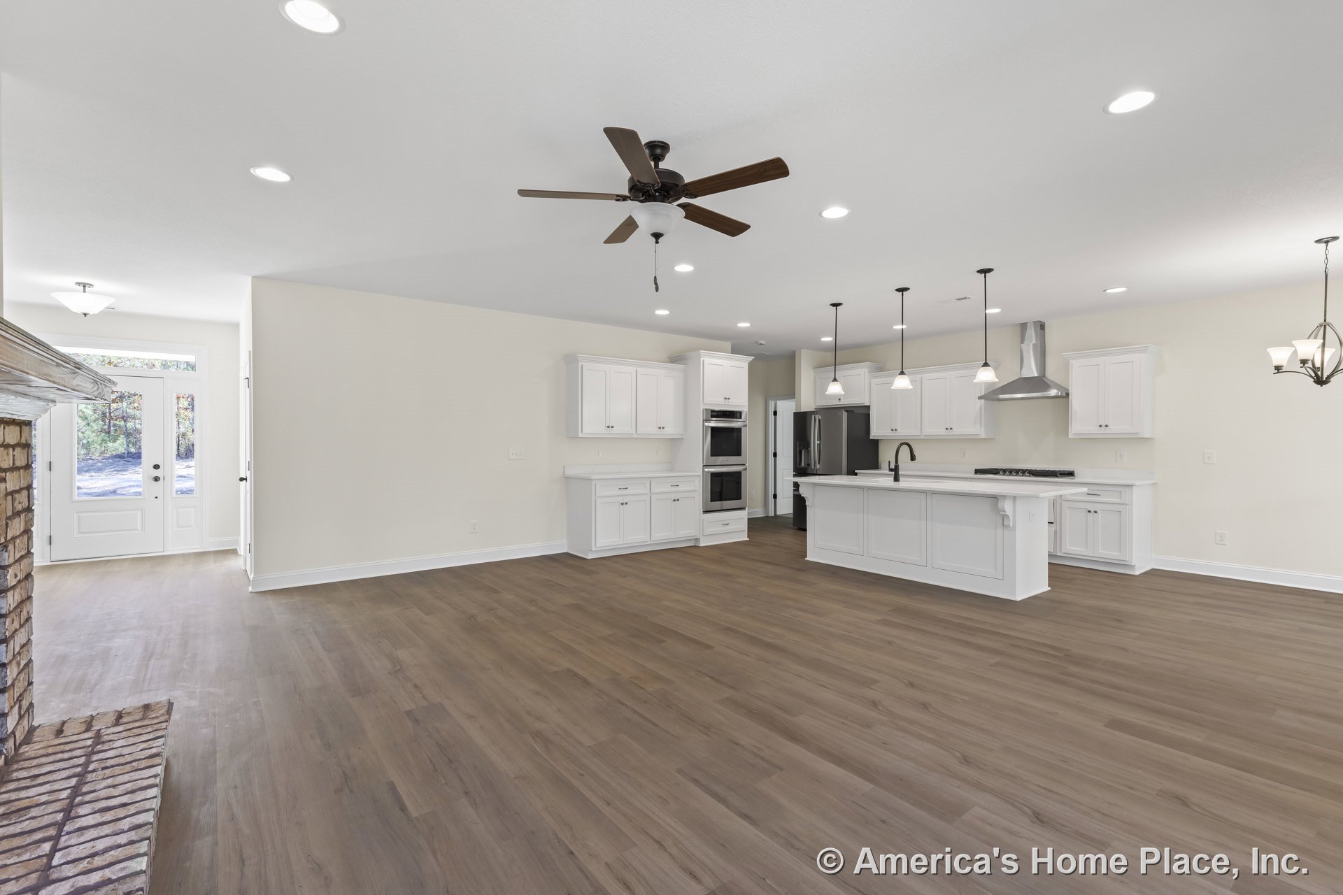 Open concept kitchen and living area with white shaker cabinets, large central island beneath pendant lights, double wall oven, stainless steel range hood, wood-look plank