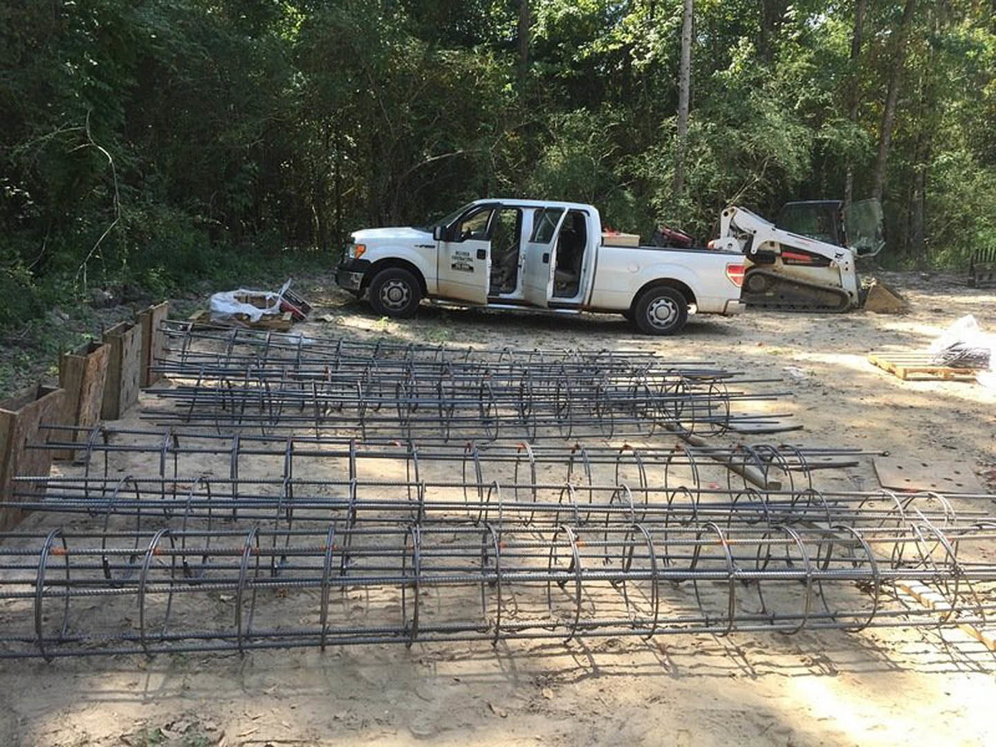 White pickup truck with open doors parked beside wooden fence, construction materials and metal rods scattered on dirt driveway, trees and partially built home exterior in