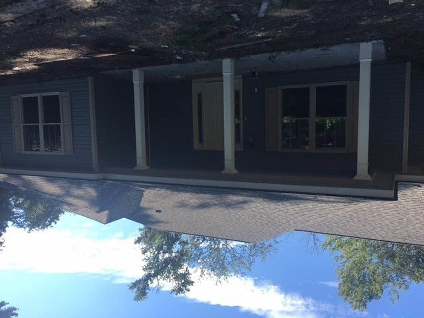 Wide front porch with white pillars, metal railing on window, tree casting shadow across sidewalk, multi-pane windows, gabled roof, leafy tree in background, partly cloudy sky