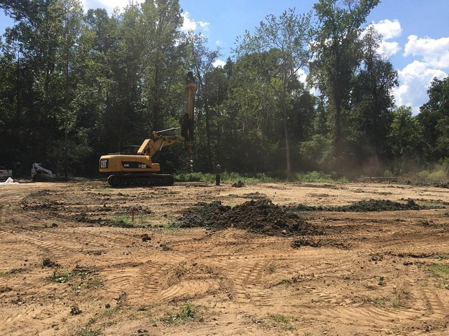 Yellow construction vehicle with extended crane arm parked on dirt lot, tire tracks and soil piles in foreground, wooded area with tall trees in background