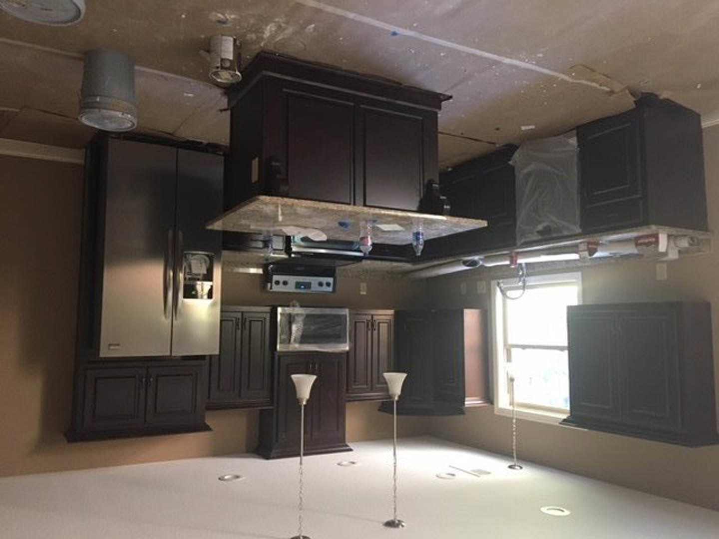 Dark wood kitchen cabinets, stainless steel refrigerator, light stone countertops, undermount sink, white walls, window with sunlight streaming in