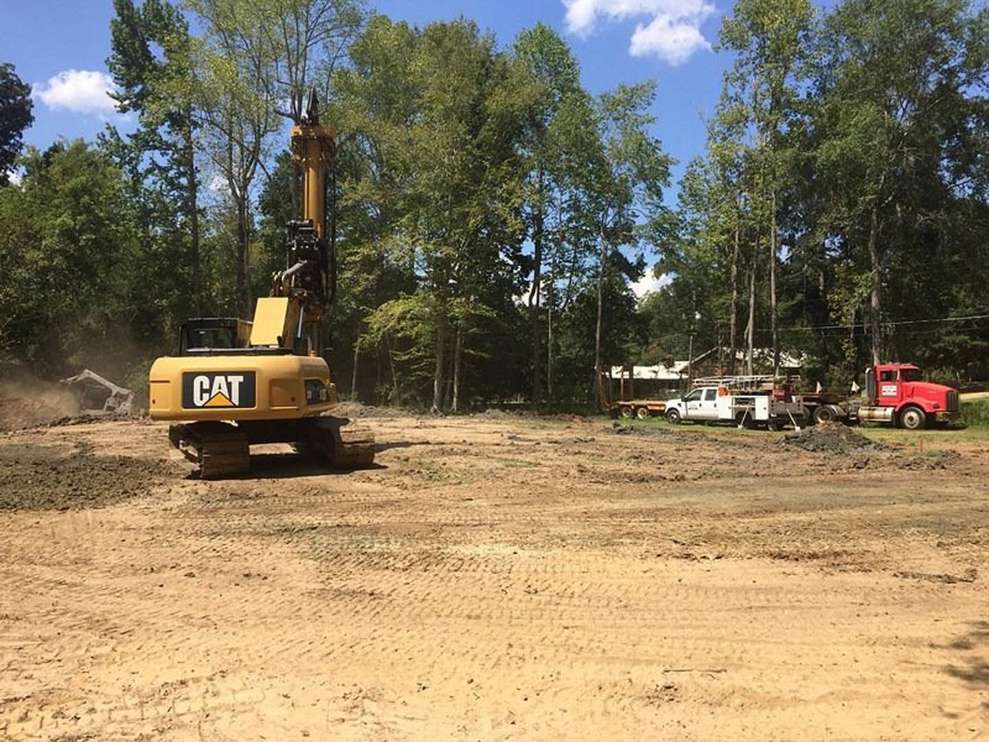 Bulldozer and white truck with ladder parked on dirt construction site, yellow and white caution sign in foreground, trees and cloudy sky in background