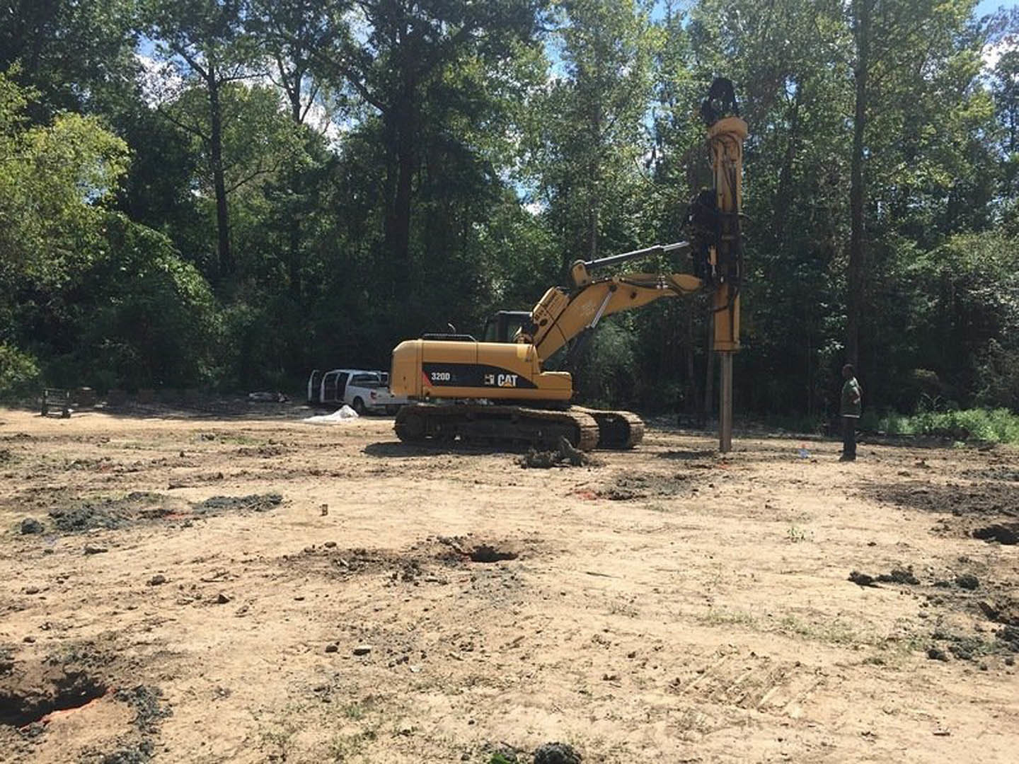 Yellow excavator with extended arm parked on dirt lot, white van with open door nearby, trees and sky in background, man standing atop construction equipment.