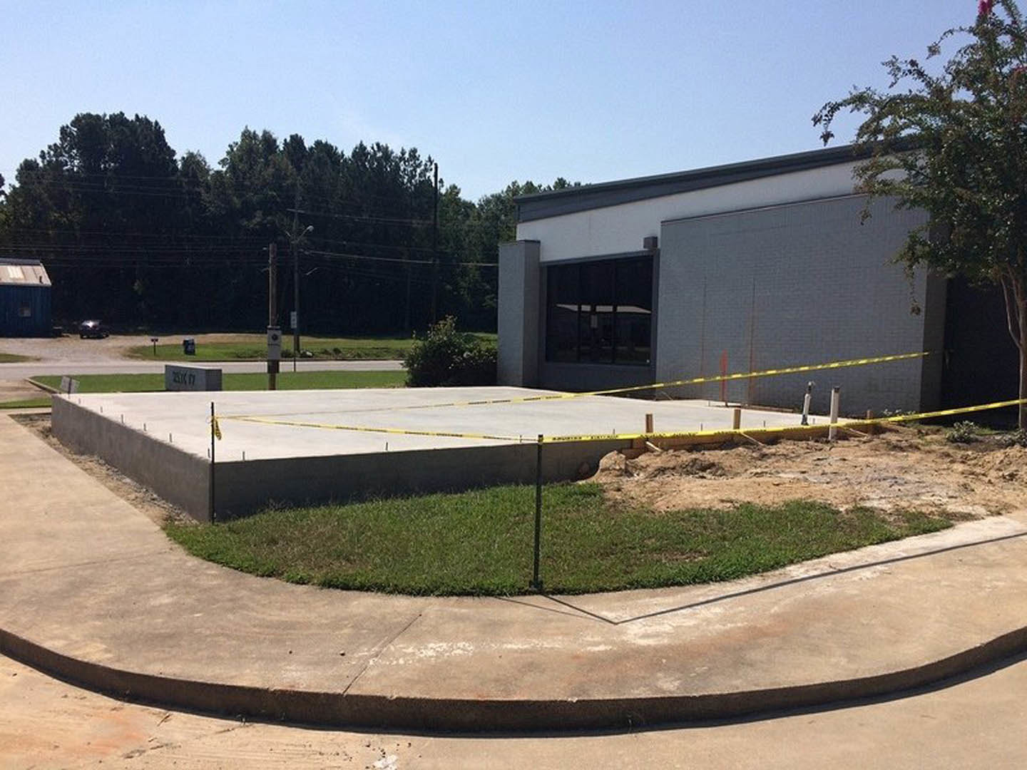 Modern residential building under construction with yellow caution tape, concrete sidewalk bordered by grass, large windows displaying signage, and mature trees alongside power