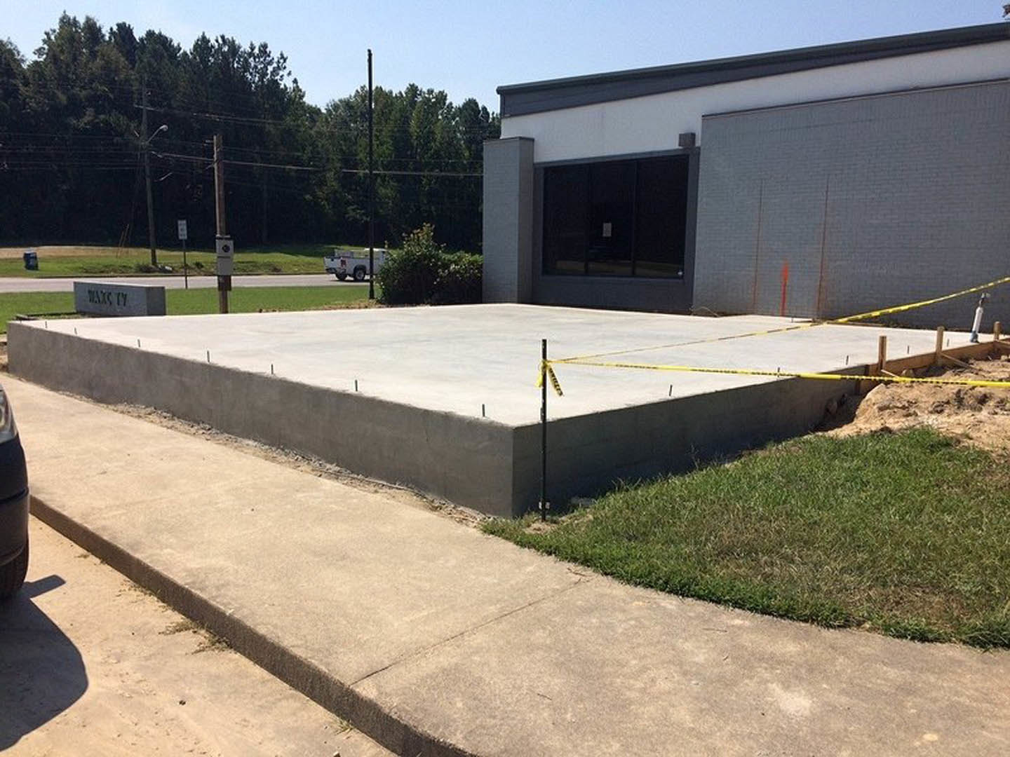 Concrete slab bordered with yellow tape, adjacent to grass patch and concrete wall, window with sign visible in background
