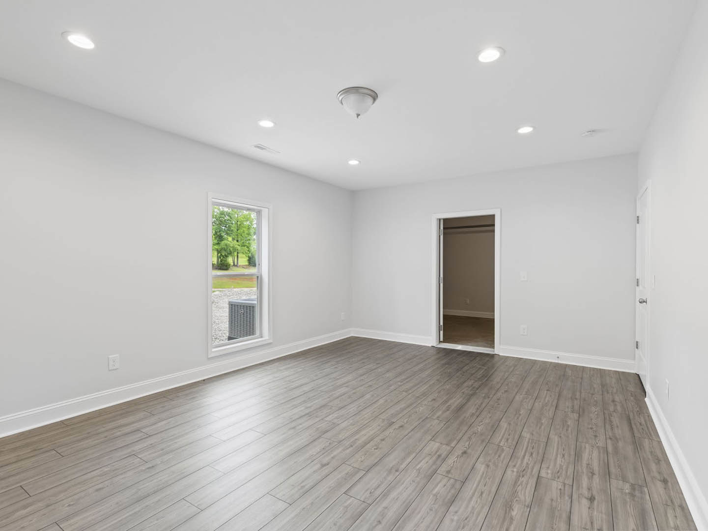 Room with light wood flooring, white walls, white door, ceiling light fixture, and window overlooking trees