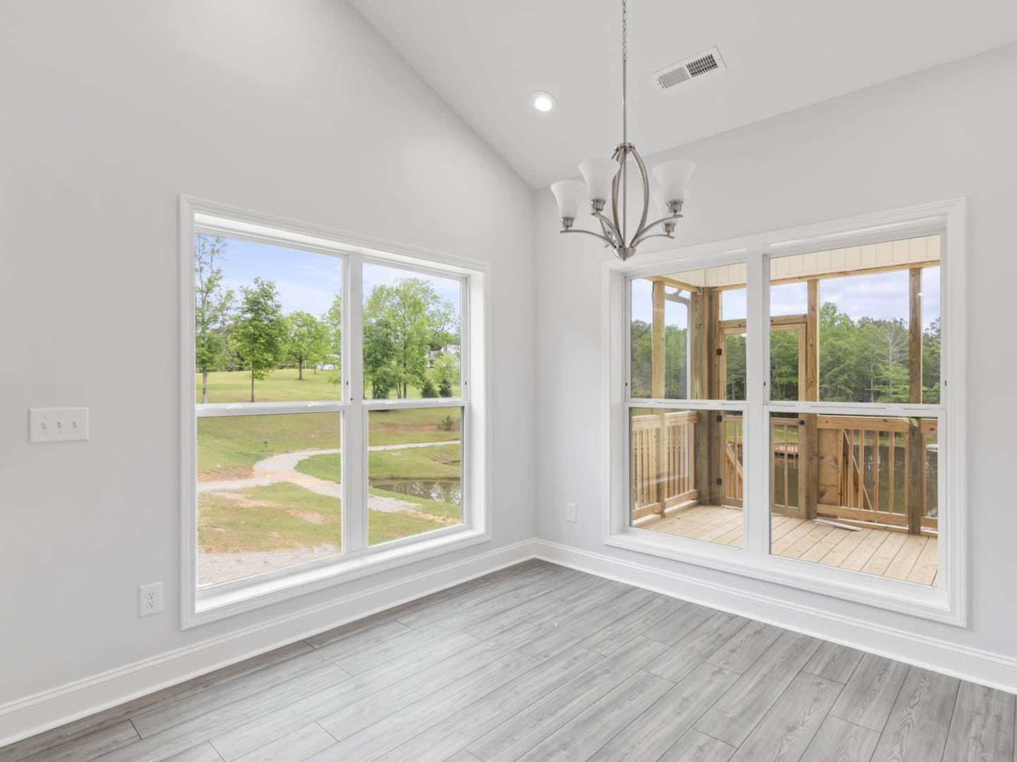 Wood-floored room featuring large windows overlooking a green lawn and trees, white wall with electrical outlet and switches, partial view of outdoor deck, close-up of modern