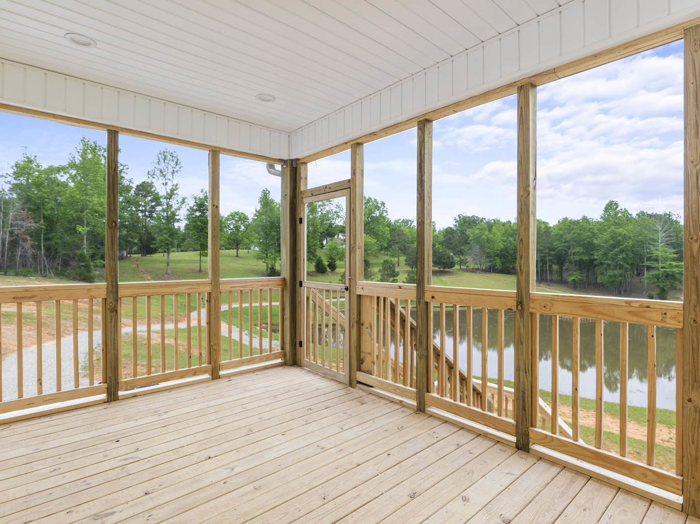 Wooden deck with white ceiling and light fixture, bordered by railings, overlooking grassy yard, lake, and surrounding trees