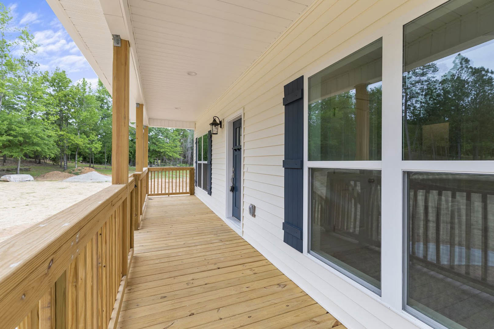 White house exterior with covered porch, wood railing, blue front door, and large windows; trees and sky in background