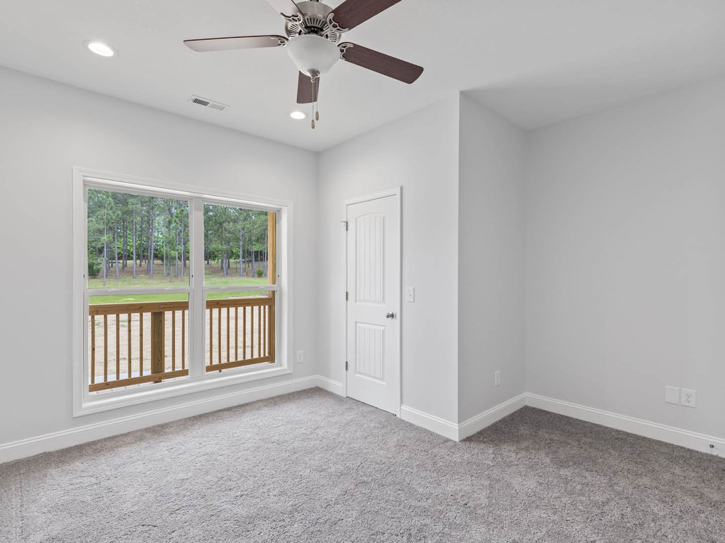 Carpeted room featuring a ceiling fan with light fixture, wooden railing, white door with silver knobs, and window overlooking trees and grass.