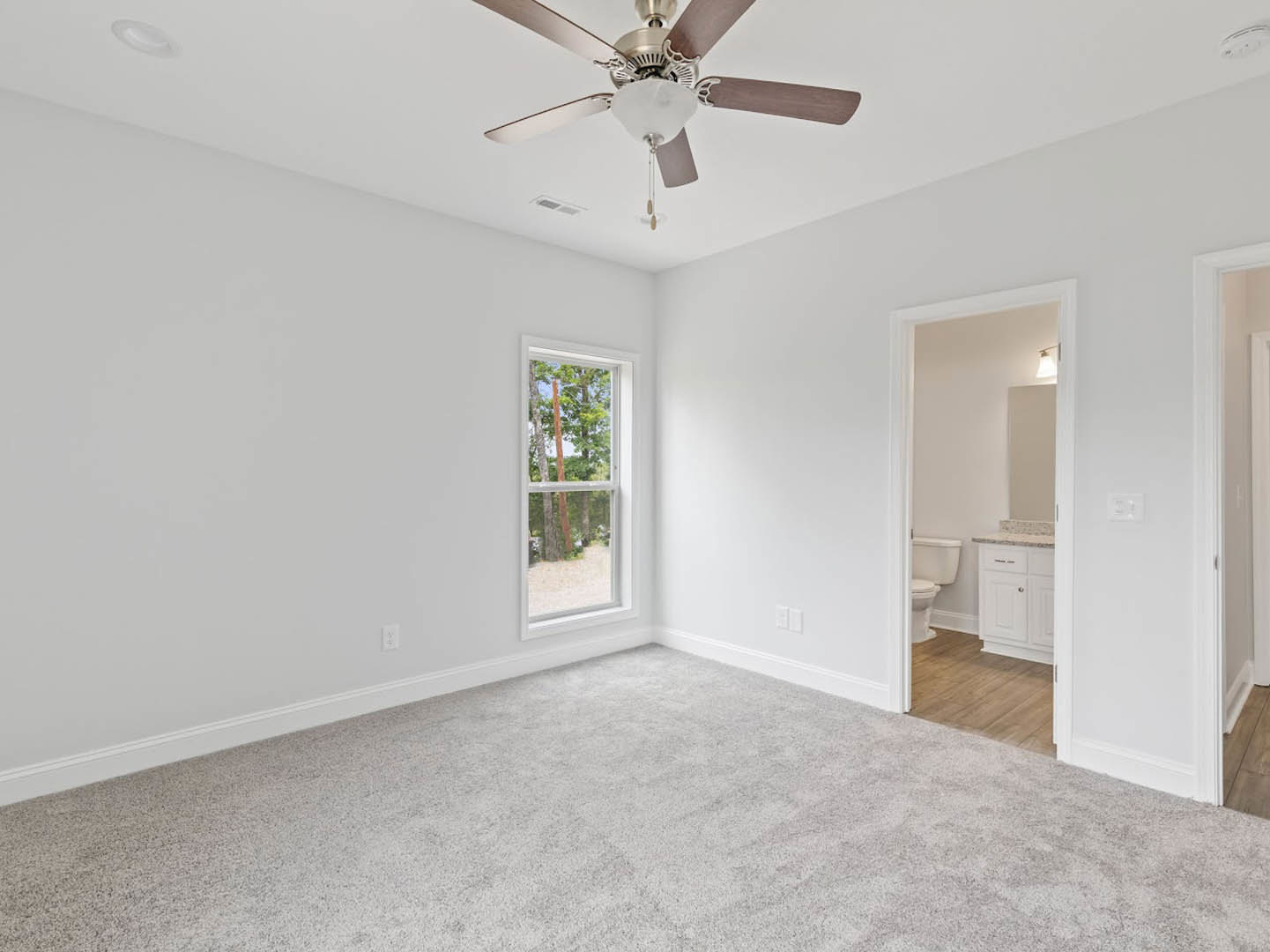 Bedroom with white carpet, ceiling fan with light fixture, large window overlooking trees, adjacent bathroom featuring white toilet