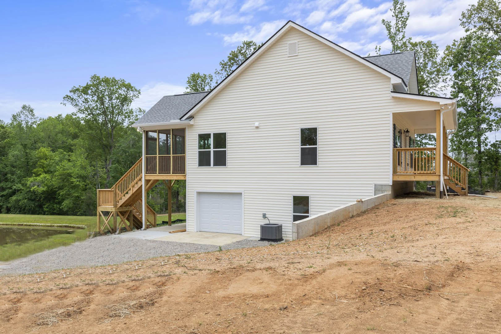 White siding house with attached garage, elevated wooden deck, stairs, and ramp leading to driveway; large window with white trim; grey rectangular utility box on ground; cloudy