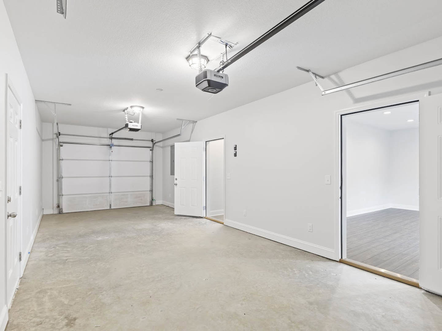 Room with white plaster walls, wood flooring, white door with silver knobs, concrete floor section, ceiling fan, and closed grey garage door.